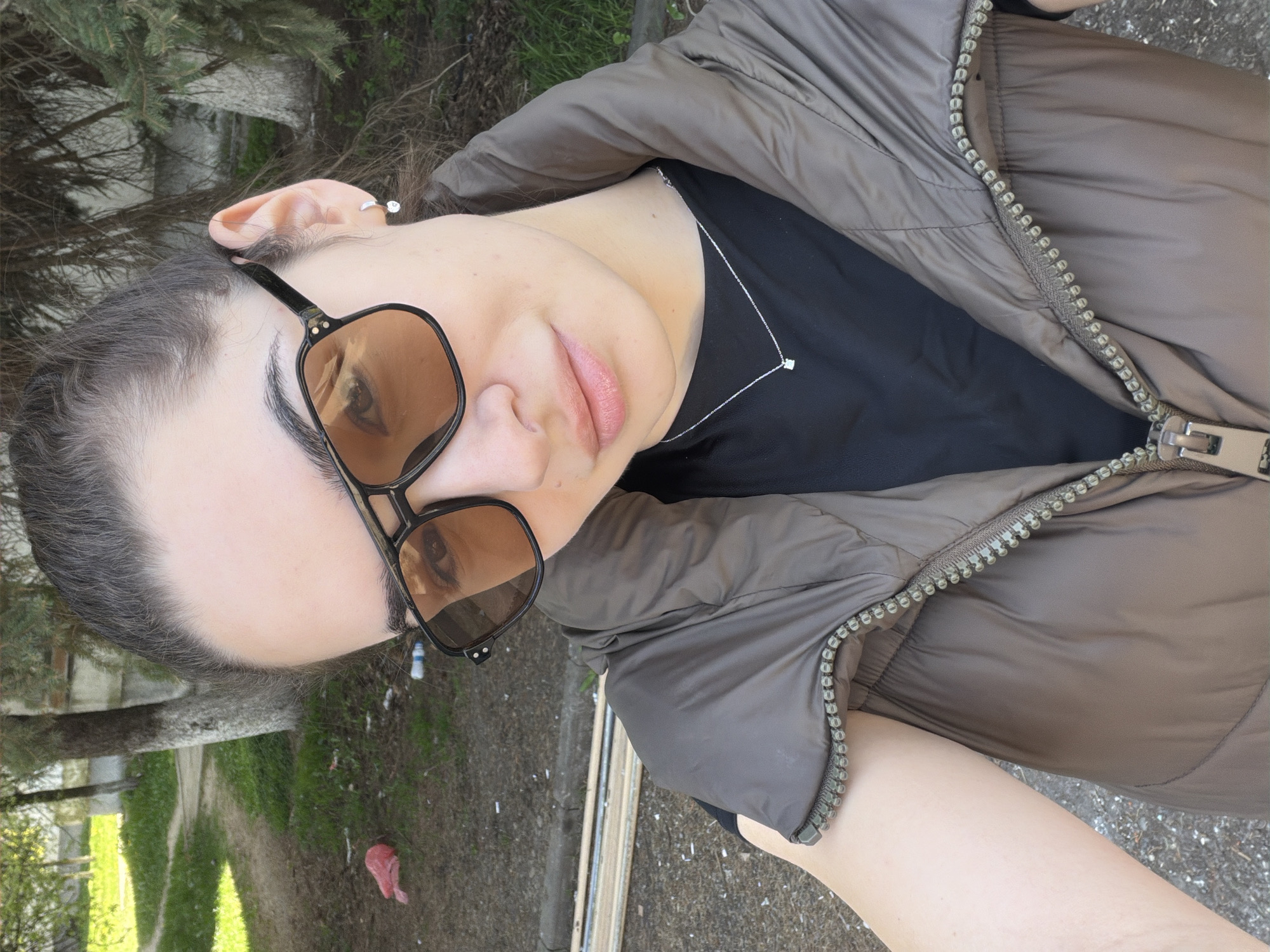 A close-up selfie captures a person from the shoulders up, tilted to the right, against an outdoor background in Sofia, Bulgaria. The subject, likely female, has dark hair pulled back and is wearing brown-tinted, square-framed sunglasses that reflect a bright light source. Her lips are slightly parted, and she has a neutral expression. A small, shiny piercing is visible in her left ear's cartilage.
She is dressed in a dark, possibly black, t-shirt under a greenish-brown puffer vest with a prominent, light-colored zipper running down the front. A delicate silver-colored necklace with a small, square pendant rests on her t-shirt.
The background suggests a park or an outdoor natural area. Behind the person, dark green foliage from trees, possibly pines, is visible. Further back, a dirt or gravel path winds through patches of green grass and more trees. The lighting appears to be daytime, casting soft shadows. On the ground in the mid-distance, a crumpled red plastic object and some white specks of debris are noticeable. No discernible text is visible in the image.