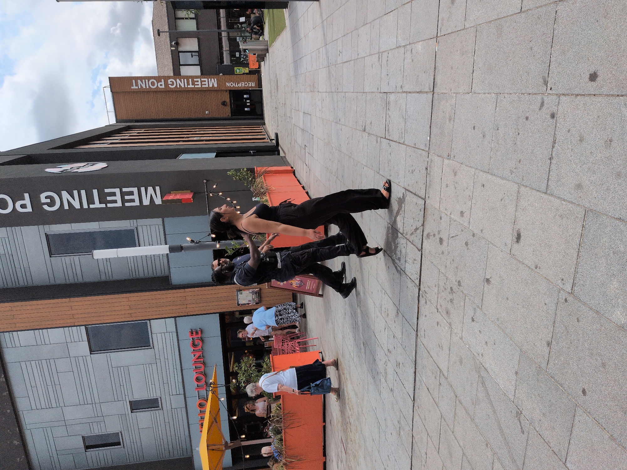The image shows an outdoor scene in Telford, United Kingdom, likely at the Meeting Point commercial complex. In the foreground, a woman in black overalls and sandals walks to the right. Behind her, a man in a dark uniform, possibly a security guard, walks to the left, with his hand near his hip. Further to the left, an older woman in a white top and black skirt walks to the left carrying a blue tote bag. In the background, people are seated at tables under a yellow umbrella outside a venue labeled "HELLO LOUNGE." The buildings feature modern architectural elements, with the "MEETING POINT" signage prominently displayed on one facade. The ground is paved with gray stone tiles, and the sky is partly cloudy.