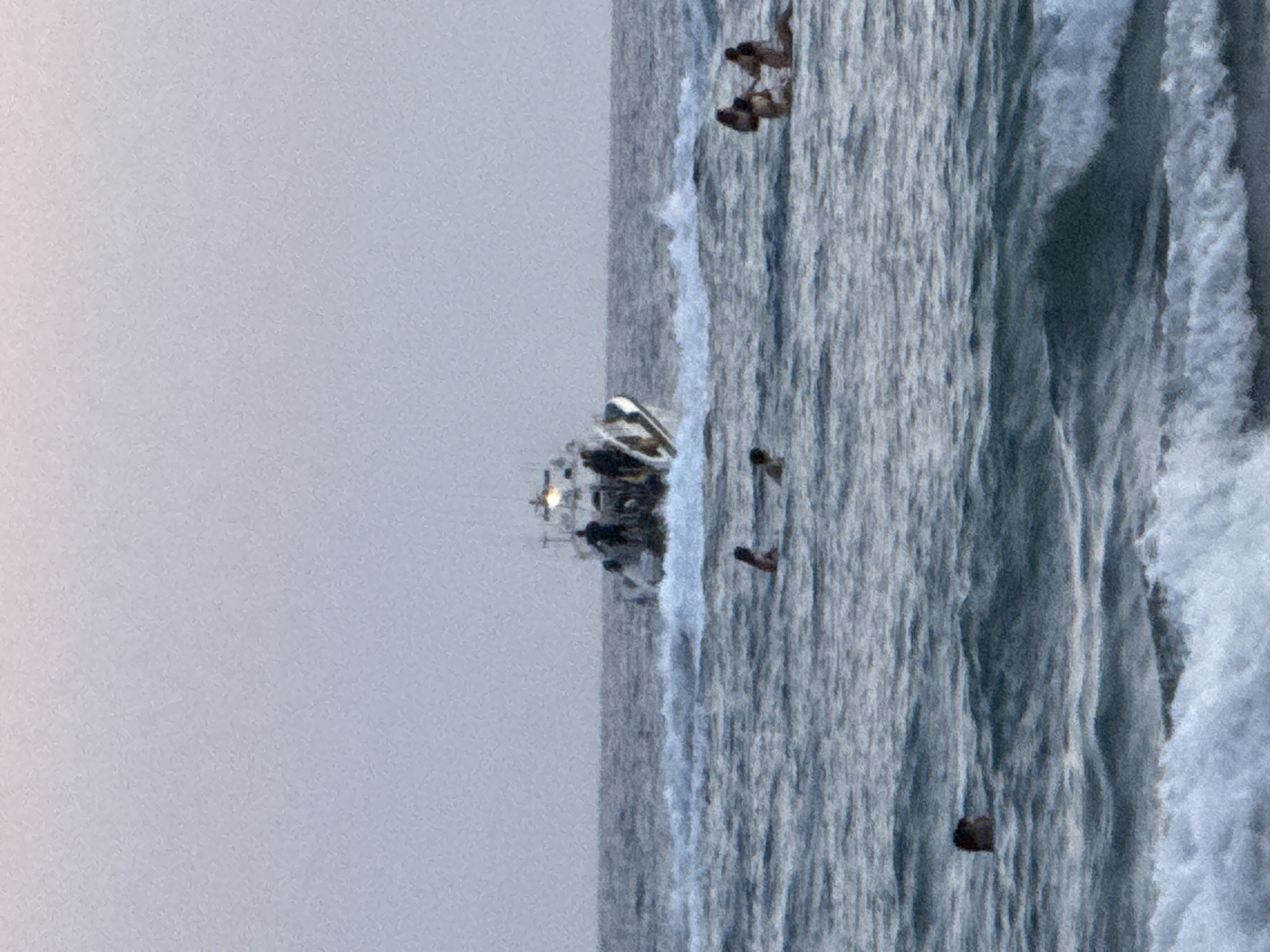 In the waters off Castelldefels, Spain, a motorboat is cruising, creating a wake. Several individuals are visible in the water. In the foreground, a person's head is just above the surface. Further out, a group of three people are swimming. To the right of them, another group of three people are partially submerged in the water. The sky is overcast and a soft pinkish-grey.