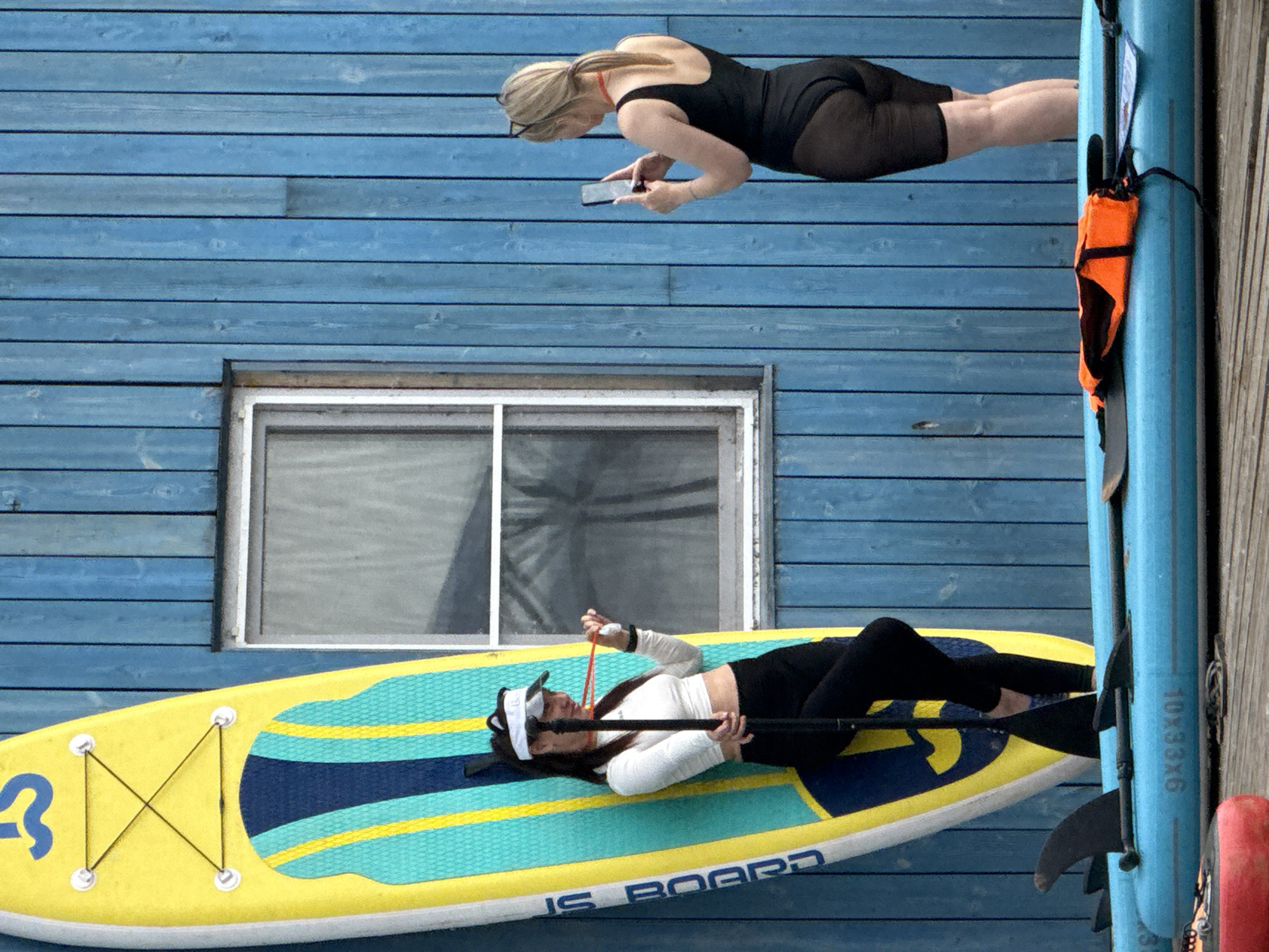 Two women are standing on a wooden dock next to a blue wooden building with a window. A yellow and blue paddleboard is leaning against the building. The woman on the left, wearing a white long-sleeved shirt and black leggings, is holding a paddle. The woman on the right, wearing a black swimsuit, is taking a picture of the other woman with her phone. Several other paddleboards are lying on the dock in the foreground, one with an orange life vest on it. The scene appears to be a recreational area possibly near a body of water.