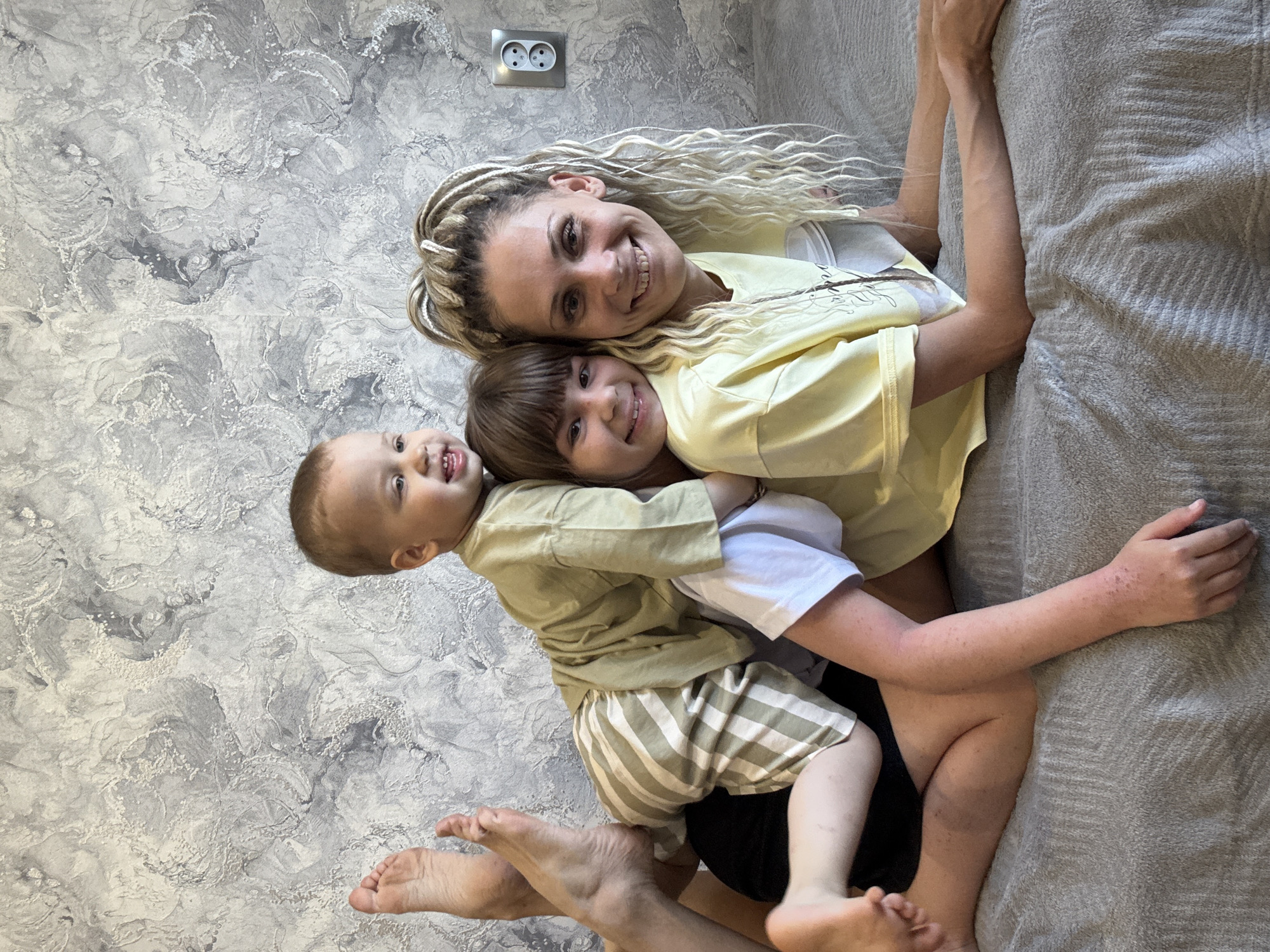 The image depicts a woman with blonde dreadlocks, wearing a yellow shirt, lying on a bed and embracing two children. One child is a toddler wearing a light brown shirt and striped shorts, and the other is a slightly older child wearing a white shirt. The background features a textured gray wall and an electrical outlet. The presence of a Russian-style outlet suggests that the location may be in Russia. The picture was taken in Rusanovo, Russia.