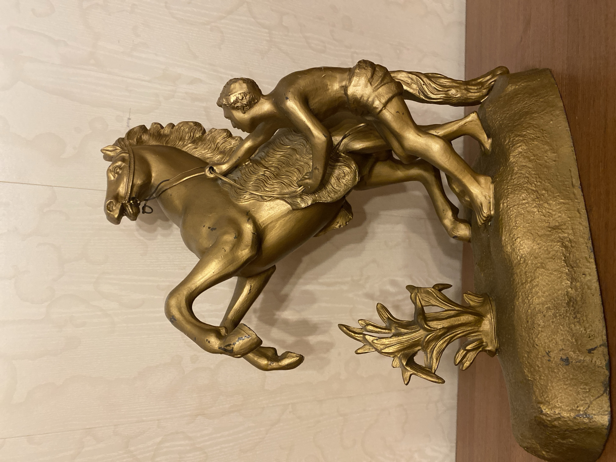 A detailed, golden-finished sculptural ensemble, approximately 18-24 inches in height, is centrally positioned on a polished, reddish-brown wooden surface, likely a table or shelf. The sculpture depicts a rearing horse with its head turned to the viewer's left, mouth slightly ajar, wearing a bridle. Its prominent mane and tail are intricately detailed. Adjacent to the horse, on its right, a barefoot, shirtless man in a loincloth is depicted. He is engaged with the horse, his right arm extended towards the horse's neck/mane, and his left arm embracing the horse's chest, suggesting an interaction of control or comfort. Both figures exhibit a metallic golden finish, showing areas of wear where darker underlying material is visible, particularly on the horse's hooves, the man's hair, and various edges of the sculpture. The figures stand on a textured, irregular, golden base. To the left of the horse on this base, a small, stylized, golden, plant-like structure with multiple upward-reaching fronds is present. The background consists of a light beige wall featuring a subtle, embossed, irregular, cloud-like pattern, with faint vertical lines also visible. The scene is illuminated by ambient light. This object is located in the city of Unknown, Unknown.