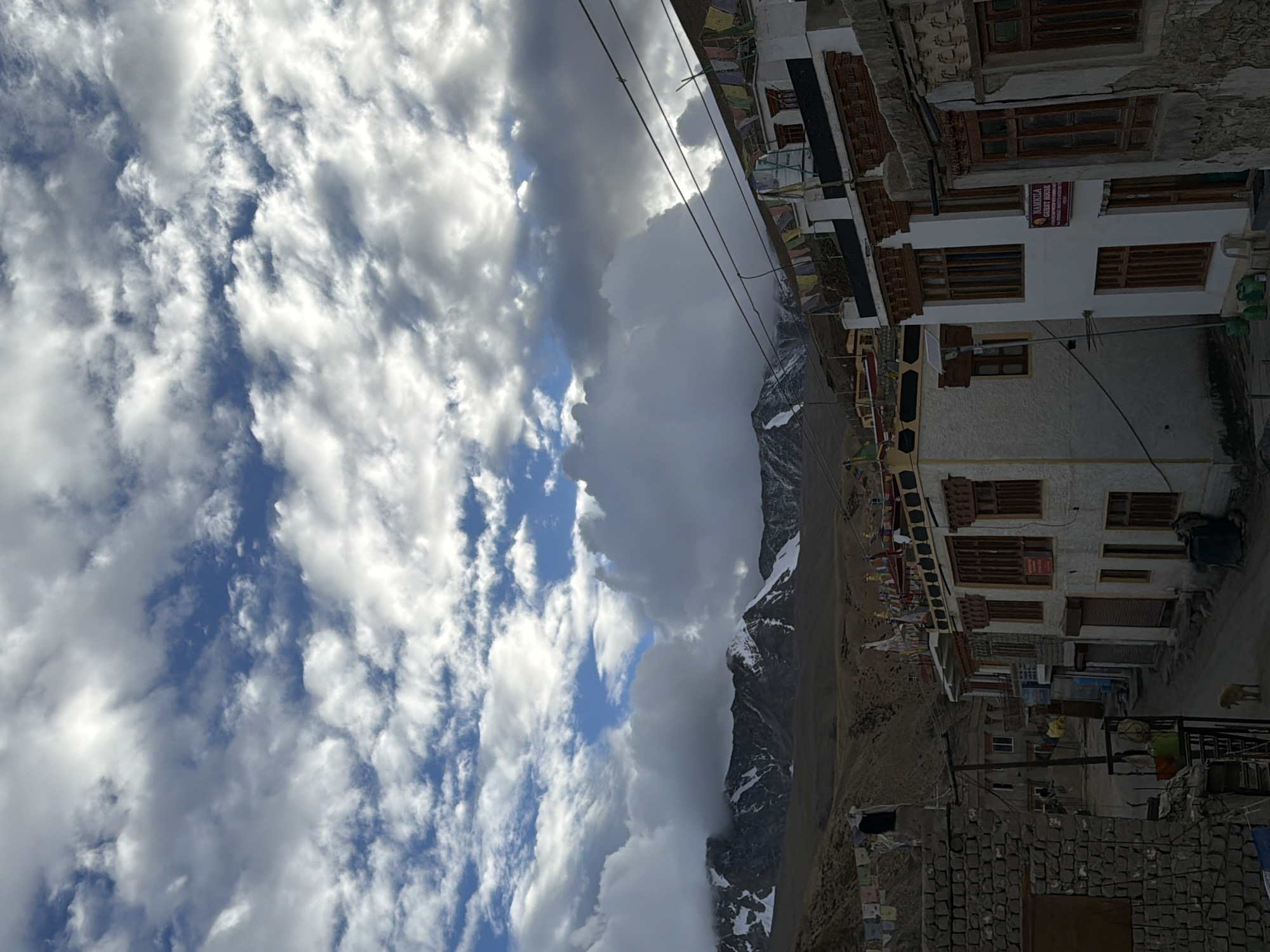 The image depicts a street scene in the village of Karzok, India, situated in the remote Spiti Valley of Himachal Pradesh. The architecture is characterized by multi-story buildings with traditional Tibetan-style flat roofs, adorned with prayer flags, a common sight in the region. The buildings are constructed with a combination of stone and whitewashed plaster, featuring wooden window frames and doors. In the foreground, a narrow unpaved road winds through the village.
The natural environment is dominated by the imposing presence of snow-capped mountains in the background, their peaks partially obscured by dramatic, cloudy skies. The mountains suggest a high-altitude, arid landscape typical of the Himalayas.
The sky is filled with a dynamic array of clouds, ranging from thin, wispy formations to dense, dramatic cumulus clouds that cast shadows over the landscape. The lighting indicates it is either early morning or late afternoon, with the sun intermittently breaking through the clouds, creating highlights and shadows.
In the lower left of the frame, a rough stone wall delineates a property. A lone dog stands on the unpaved road in the center, appearing to be unattended. Prayer flags are strung across wires and rooftops, fluttering in the breeze, adding a splash of color to the scene. One building prominently displays a sign reading "HAMKILA GUEST HOUSE". The overall atmosphere conveys a sense of peaceful isolation and the rugged beauty of a Himalayan village.