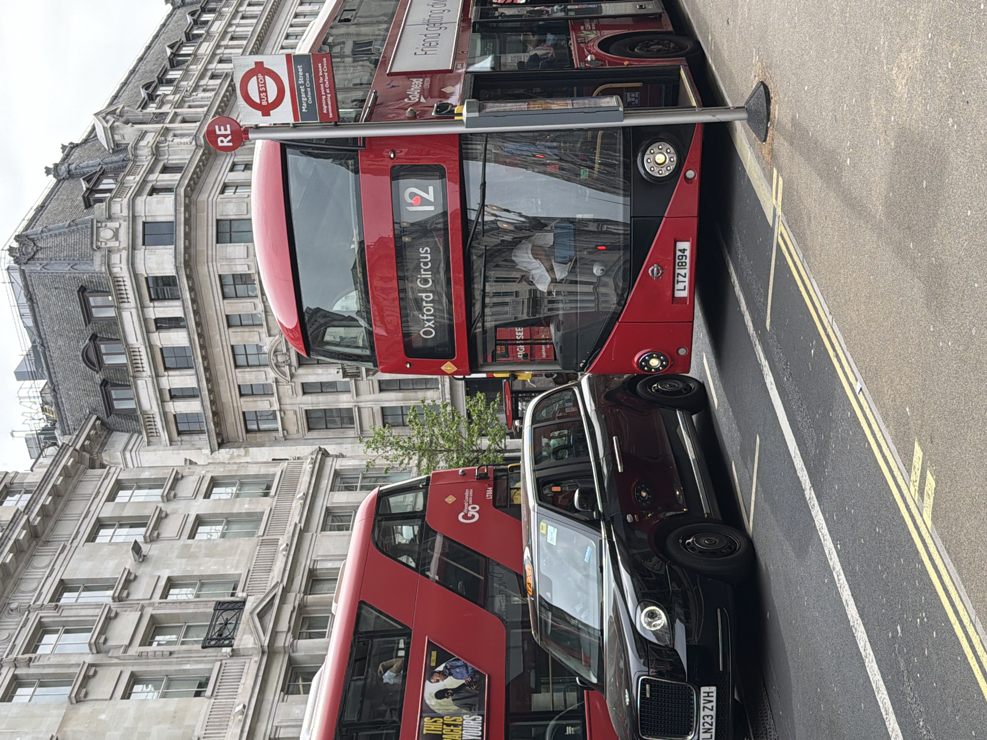 The image captures a street scene in London, United Kingdom. In the foreground, a black taxi is parked alongside a red double-decker bus. The bus is numbered 12 and its destination is Oxford Circus. The bus has license plate LTZ 1894. The taxi's license plate is LN23 ZVH. A bus stop sign is visible, indicating it is on Margaret Street, Oxford Circus, and serves as an alighting point for buses terminating at Oxford Circus. The background shows the architecture of a tall building, with rows of windows. An advertisement on the lower level of the other bus reads "THIS ACE IS YOURS".