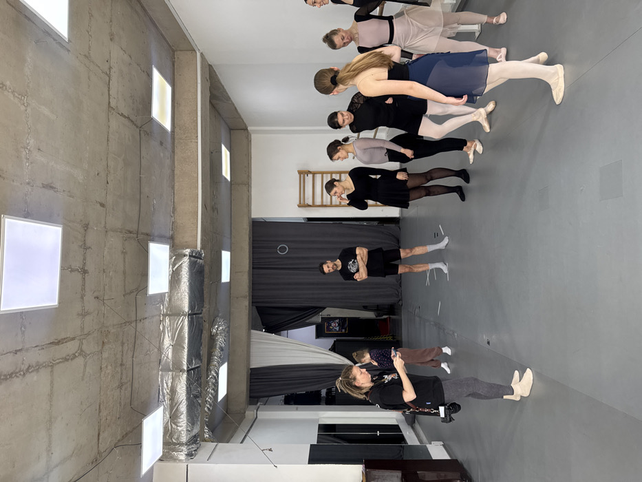 An indoor scene depicts a large room, likely a dance or ballet studio, located in Unknown, Unknown. The room features a grey floor, white walls, and an exposed concrete ceiling with visible conduits and multiple rectangular LED panel lights. Silver foil-wrapped ductwork runs across the ceiling. Mirrored panels are partially visible on the left wall, and a wooden wall-mounted barre is positioned on the back right wall. Dark grey and white curtains are visible at the rear of the room, suggesting entrances or changing areas.
Ten individuals are present. In the foreground left, a female with light brown hair, wearing a black t-shirt, grey leggings, and light-colored foot coverings, holds and observes a smartphone. A black camera hangs from a strap across her body. Behind her, a young female child, dressed in a brown long-sleeved top and brown pants with white socks, stands looking downwards.
In the mid-ground center, a male individual with a beard, wearing a black t-shirt and black shorts, stands with his arms crossed, facing right. He wears white socks.
To the right, a line of seven female individuals, primarily young adults, are positioned. They are all dressed in various forms of ballet or dance attire, including black leotards, sheer wrap skirts (in dark blue and light pink/nude), black shorts, light grey long-sleeved tops, tights, and light-colored ballet shoes or socks. Their postures vary, with some looking forward, others looking towards individuals in the group, and one touching her ear. A ballet barre is partially visible along the far right wall behind them. The individuals appear to be awaiting instruction or observing the scene.