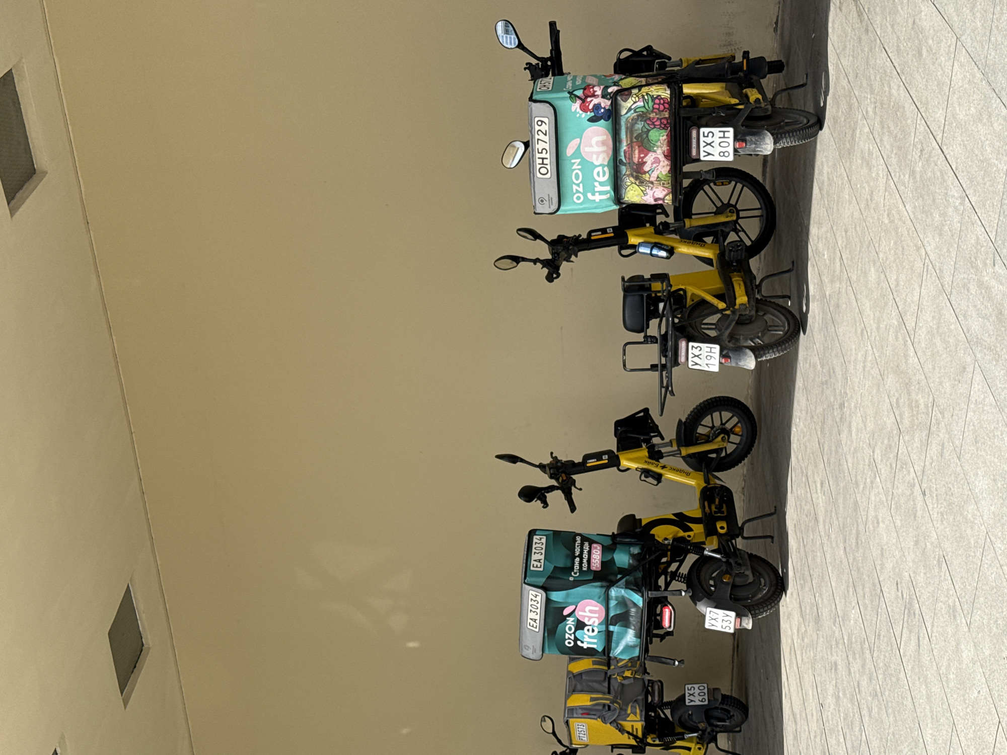 The image shows four yellow delivery scooters parked in a row against a light-colored wall. Two scooters are on the left, and two are on the right. Each scooter has a large, insulated delivery box on the back. The boxes on the left scooters are light blue and green with "OZON fresh" branding. The boxes on the right scooters also have "OZON fresh" branding and are decorated with fruit images. The scooters are parked on a light-colored tiled ground. The wall behind the scooters has a subtle shadow pattern. There are no people visible in the image. The license plates on the scooters are visible, displaying alphanumeric characters such as "EA 3034", "YX5 600", "YX7 53Y", "YX3 19H", "OH5729", and "YX5 80H". The branding on one of the scooters includes "Яндекс.Еда", which translates to "Yandex.Eda," a Russian food delivery service. This suggests the location is likely in Russia, and given the prevalent use of Yandex services, potentially Moscow.