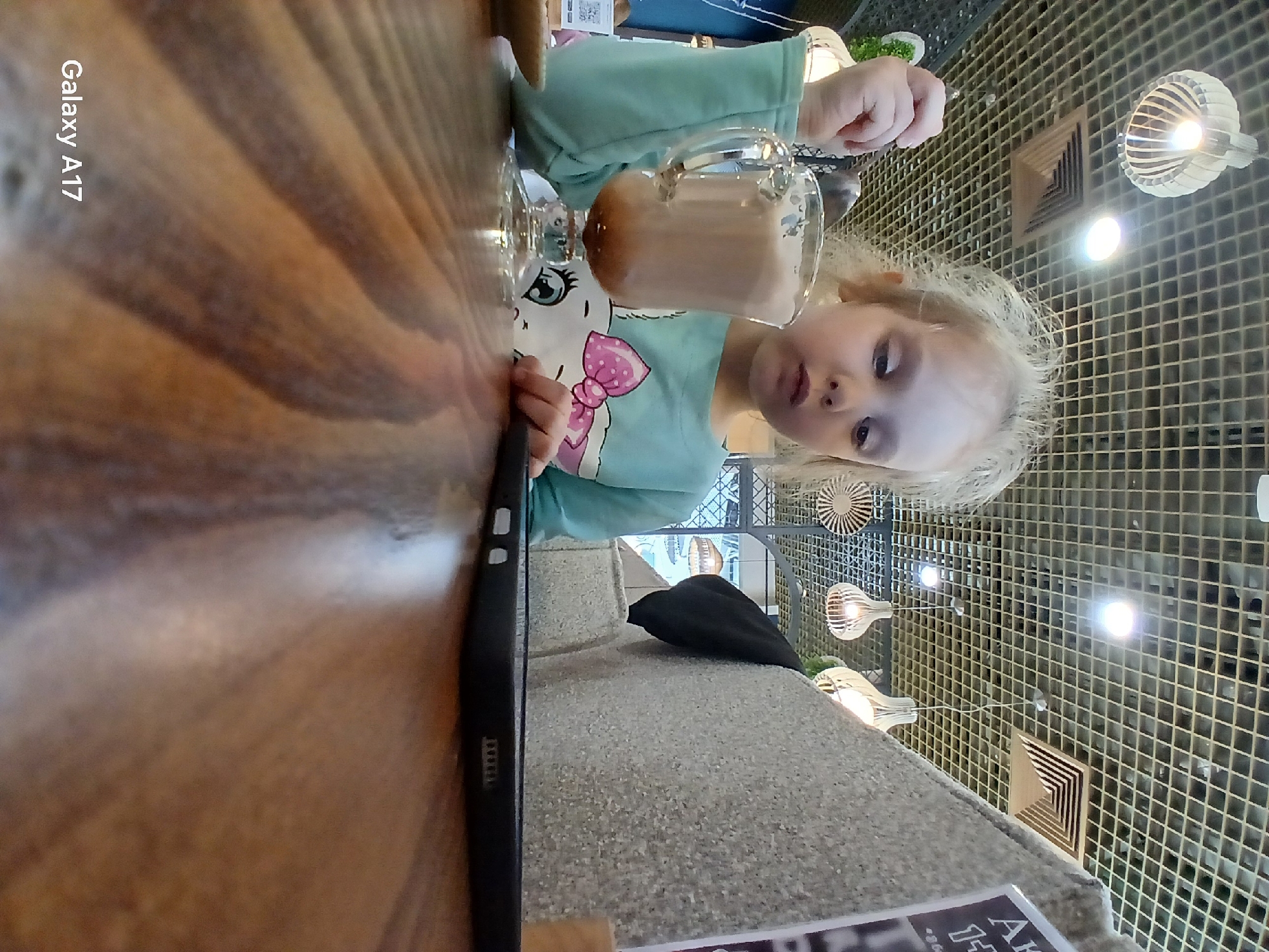A young Caucasian child with blonde, disheveled hair, appears seated at a wooden table in a modern cafe within a shopping center in Yekaterinburg, Russia. The child, wearing a light turquoise long-sleeved shirt adorned with a cartoon character featuring a pink bow, holds a spoon immersed in a clear glass mug containing a frothy, light brown beverage with a darker layer at the bottom. Her gaze is directed downwards, possibly towards the drink or a black tablet/smartphone resting face-down in a case on the table to her right. The table exhibits a prominent wood grain. The background features a grey, textured, upholstered booth or seating. The ceiling is a grid system with integrated lighting and multiple modern, decorative pendant lights with a slatted wooden design. A square air vent is also visible in the ceiling. To the far left, a small, light-colored dish and a stand with a QR code are partially visible. A partial promotional sign with Cyrillic text and the number "14" is seen in the upper right background.