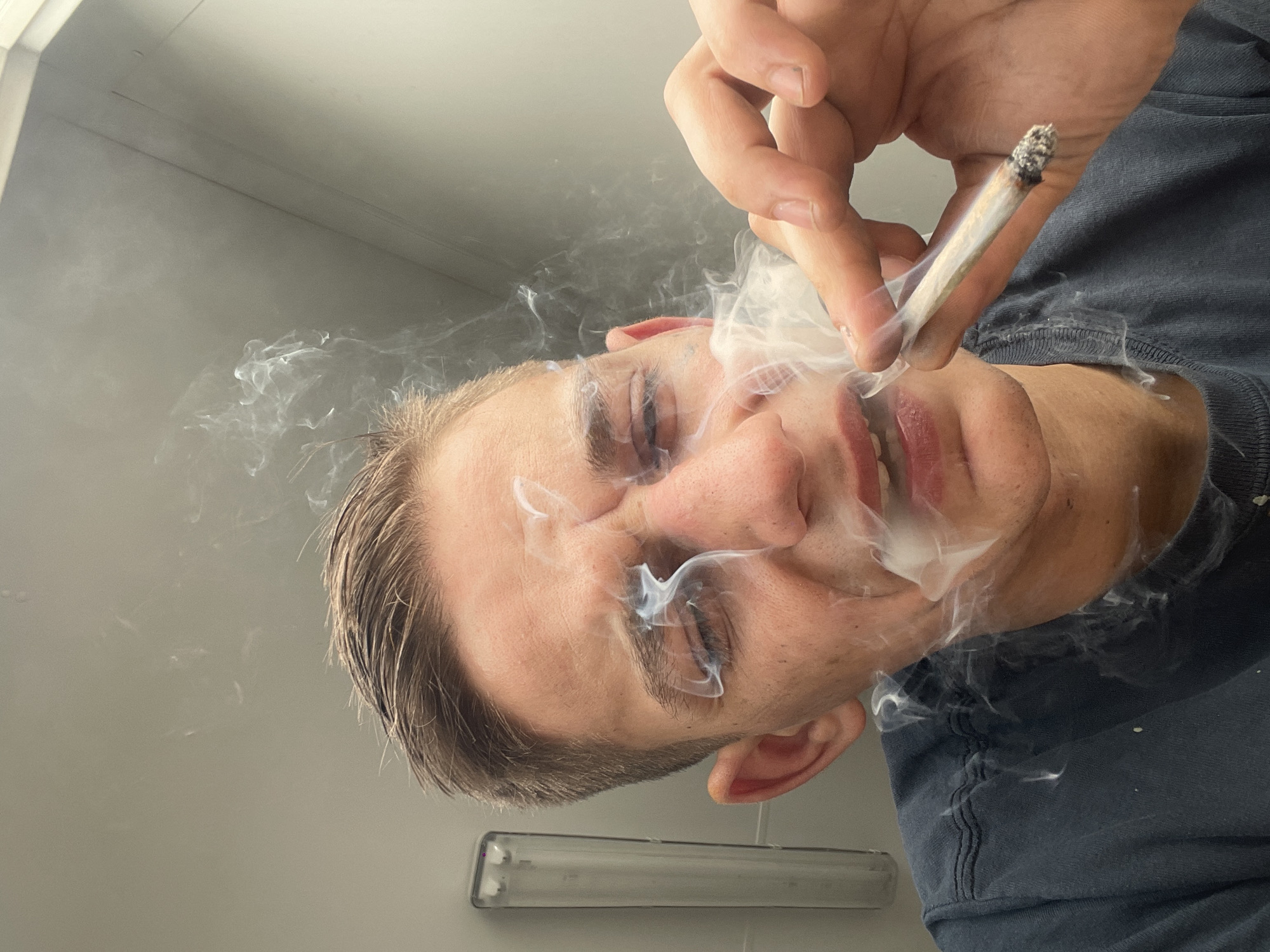 A man is holding a lit cigarette or joint in his right hand, exhaling smoke upwards towards the camera. He is indoors, possibly in a bathroom, judging by the fluorescent light fixture visible in the lower part of the frame. He is wearing a dark-colored shirt. His head is tilted to the side. The location is Unknown, Unknown.