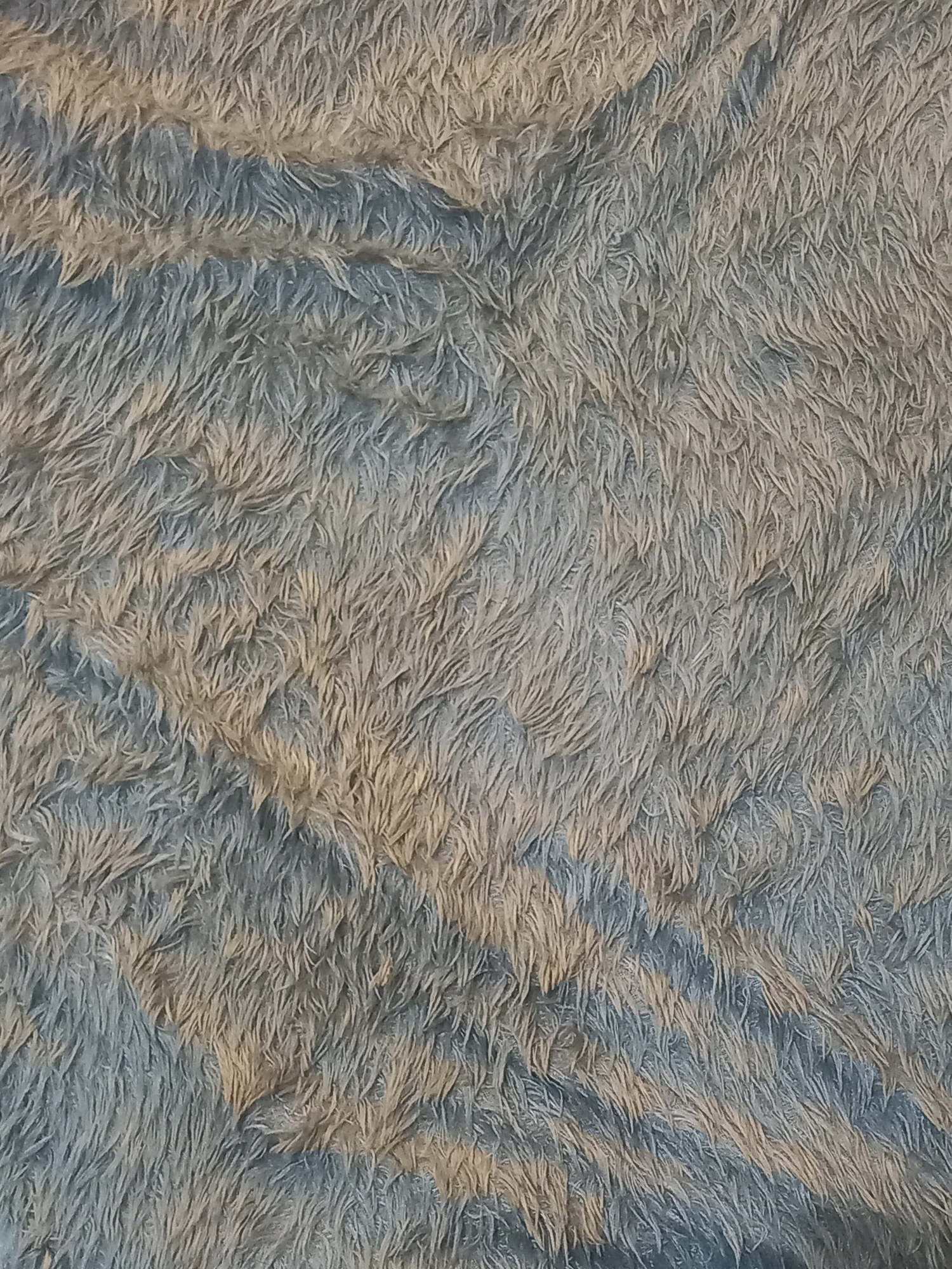 The image displays a close-up, top-down view of a textured surface, likely a rug or blanket. The material is fluffy and appears to have a shag or faux fur texture, with individual fibers clearly visible. The color palette consists of muted shades of light blue and a sandy beige or light brown, creating a marbled or abstract pattern. The fibers are short and densely packed, with variations in direction contributing to a sense of organic flow and texture. There are no people, objects, or actions depicted. The lighting creates subtle shadows, highlighting the depth and texture of the material. The specific location in Bendo, Indonesia, is not discernible from the visual information provided in the image.