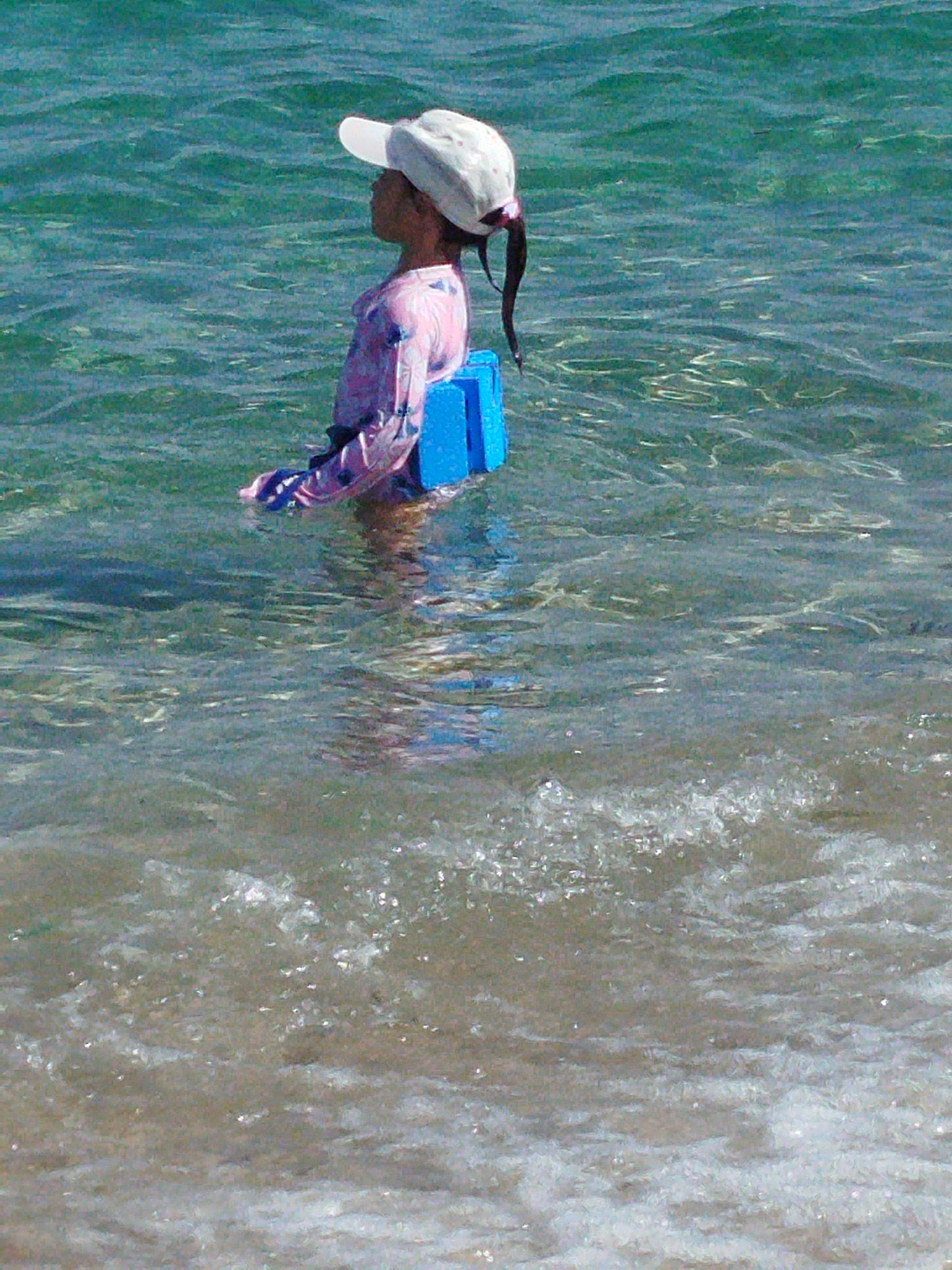 In Sithonia Municipal Unit, Greece, a young child wearing a white hat and a pink long-sleeved shirt is sitting in shallow, clear turquoise water. The child is partially submerged, with two bright blue flotation devices positioned behind their back, appearing to support them. The water is rippling and reflecting sunlight. The background shows more of the wavy sea. In the foreground, gentle waves are lapping at a sandy beach. The image captures a peaceful moment of a child enjoying the water.