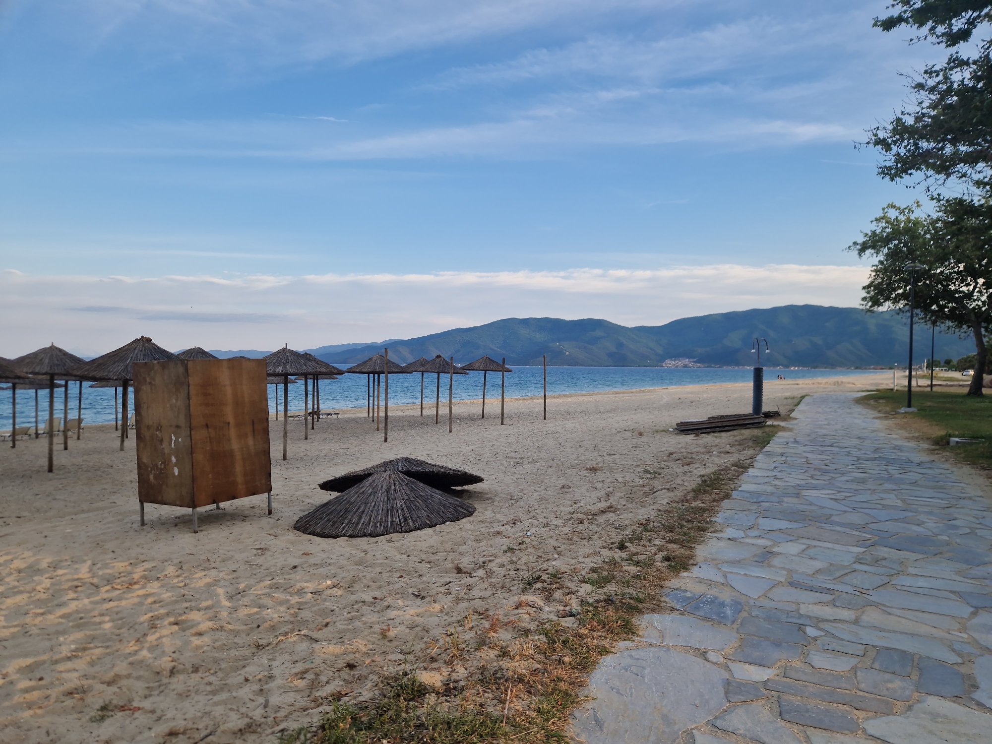 The image captures a serene coastal scene in Ασπροβάλτα, Greece. The foreground is dominated by a sandy beach, dotted with straw parasols, some standing upright and others collapsed. A rectangular, wooden structure is positioned near the parasols. A stone-paved pathway runs along the right side of the image, lined with trees and lampposts. In the background, a calm sea meets a mountain range under a partly cloudy, blue sky. Two people are visible in the distance.