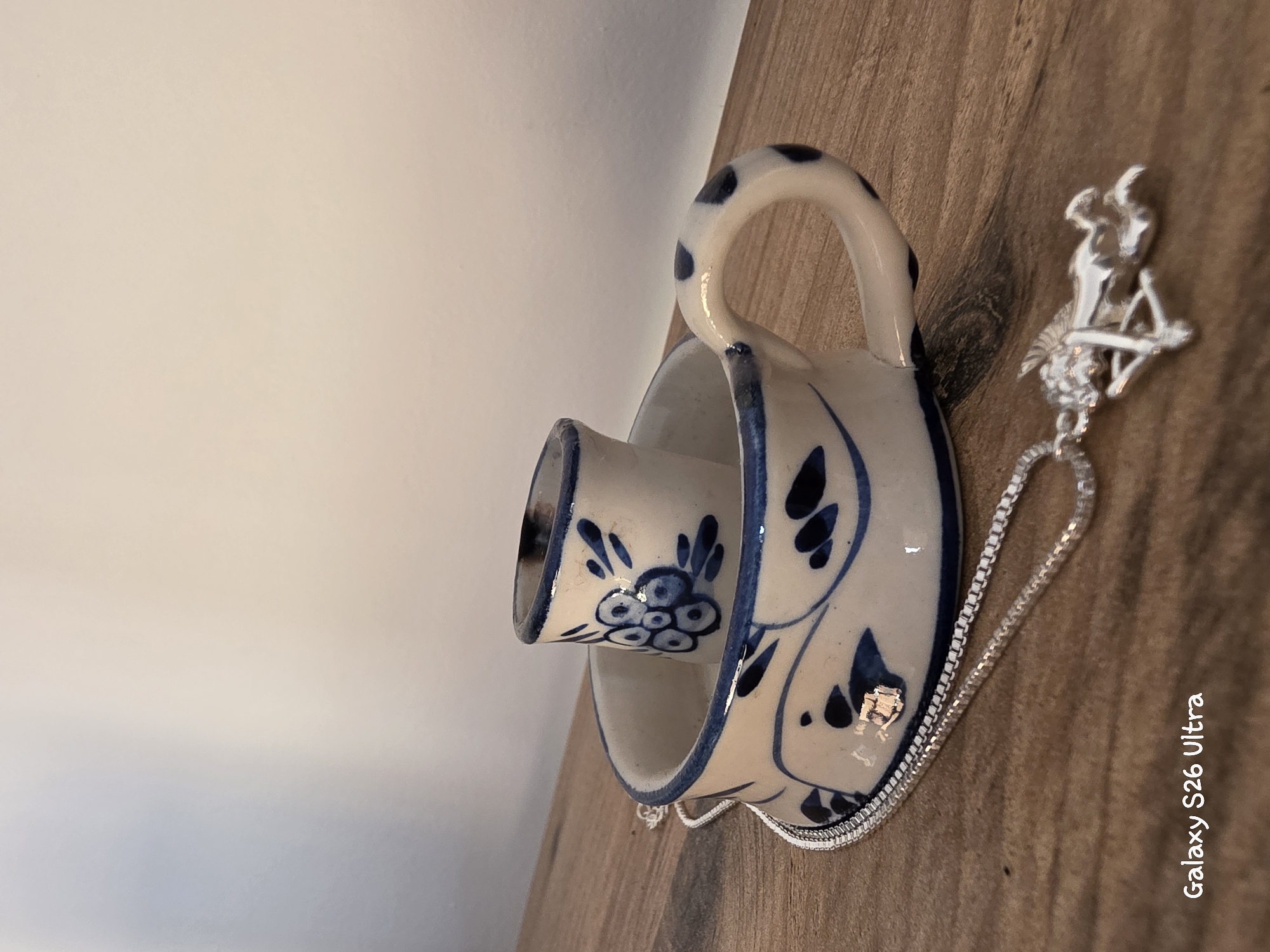 A silver necklace with a pendant of a winged mythical creature rests on a wooden surface, partially in front of a decorative ceramic cup with blue floral patterns. The cup appears to be a candlestick holder, with a smaller, cylindrical piece placed inside the larger cup. The background is a plain, light-colored wall. The image includes a watermark in the bottom left corner that reads "Galaxy S26 Ultra". There are no people present in the image, and no specific location in Plovdiv, Bulgaria, can be identified.