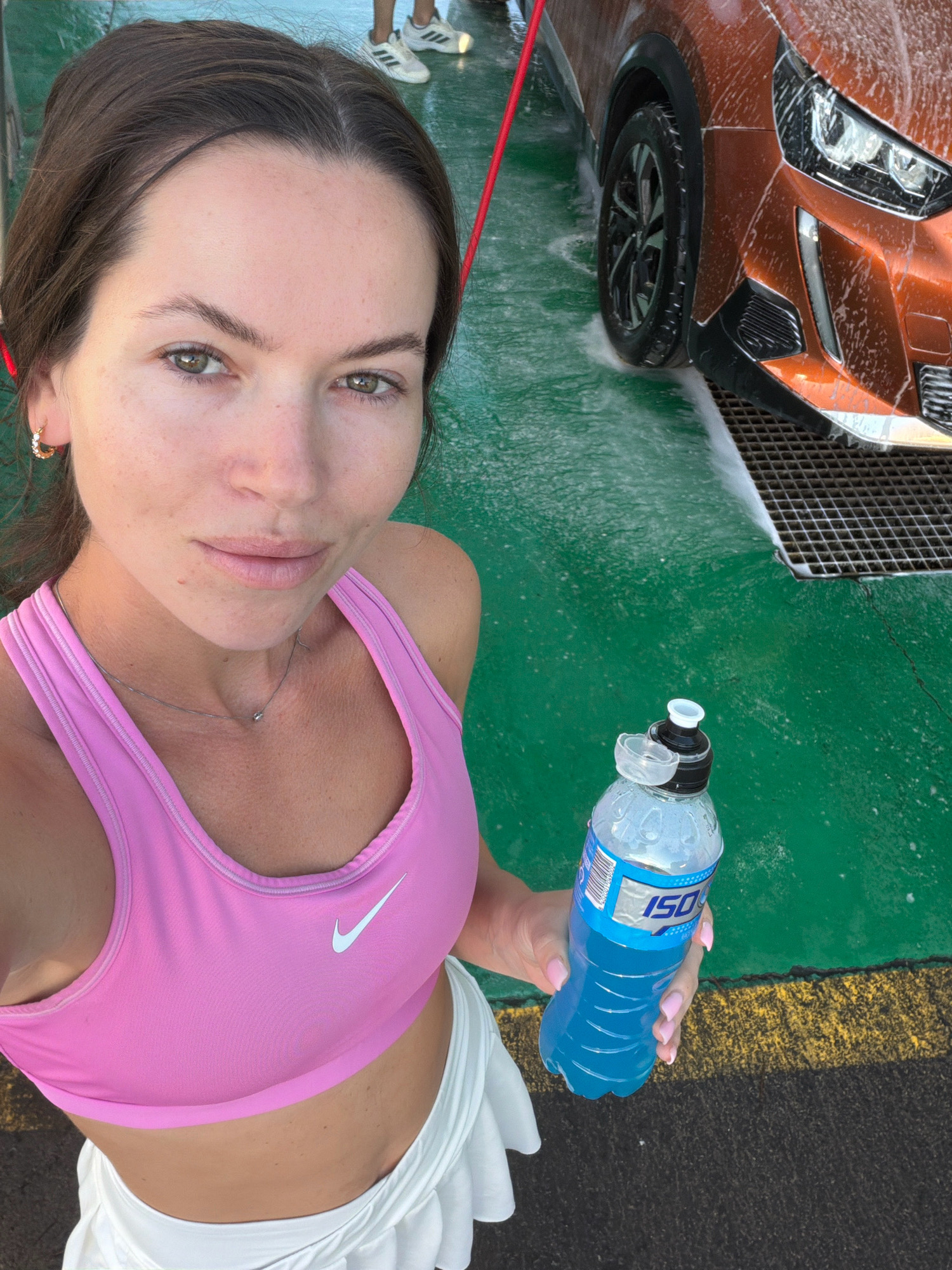 In Cambrils, Spain, a woman with brown hair, wearing a pink Nike sports bra and a white skirt, is taking a selfie while holding a blue sports drink. She has light skin, green eyes, and is wearing a thin silver necklace and gold hoop earrings. Behind her, an orange car is being washed, with soap suds and water on the green ground. The car has a black tire and chrome accents. The scene appears to be an outdoor car wash.