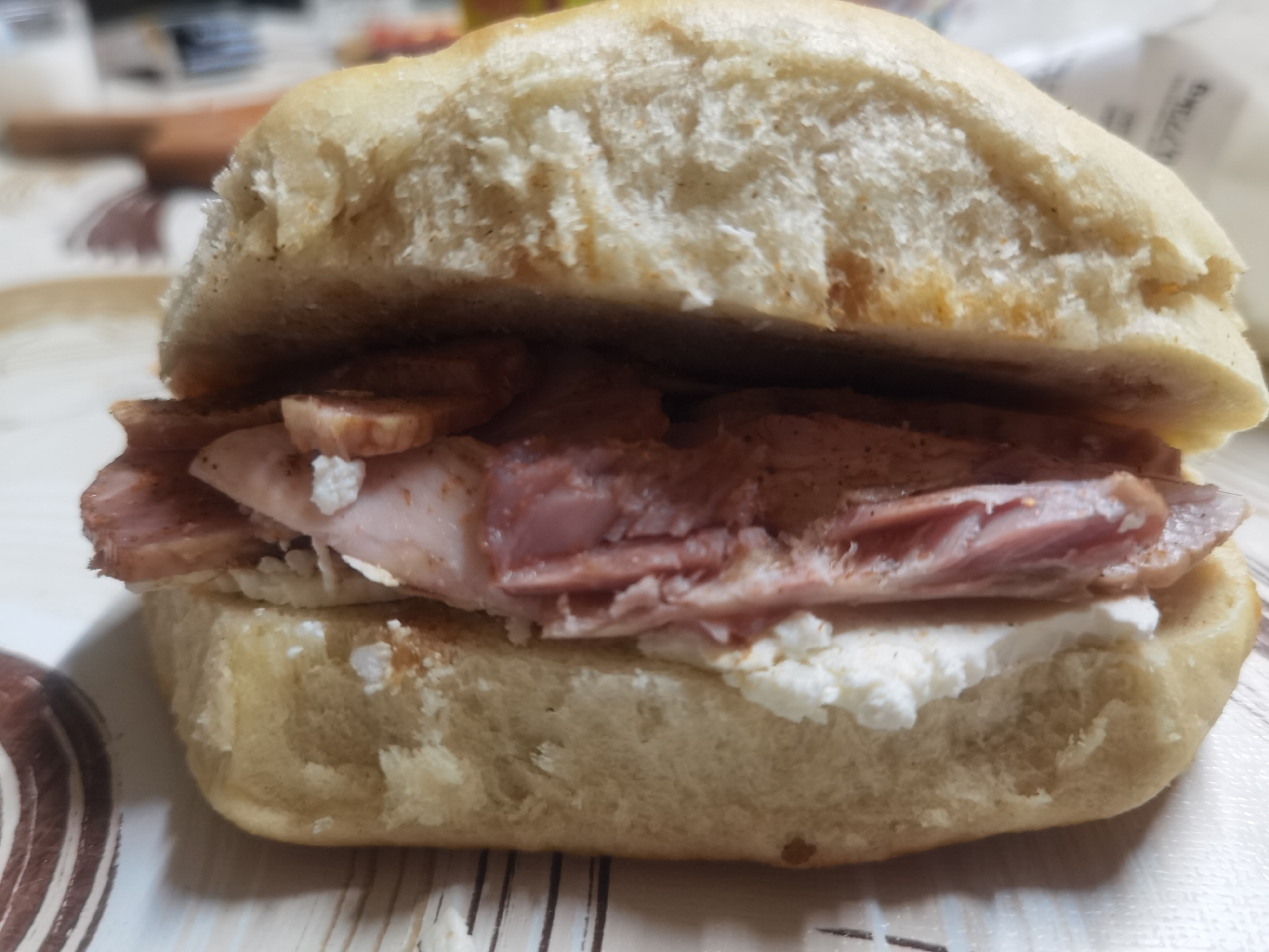 A close-up, eye-level photograph depicts a sandwich positioned centrally on a patterned surface. The sandwich consists of a light-colored, soft bread bun, horizontally sliced. The upper half of the bun covers a filling composed of multiple thin, reddish-pink to brownish-pink slices of cooked meat, consistent with ham or a similar pork product, with some browning visible on the edges of the meat. Below the meat, a layer of white, crumbly substance, indicative of soft cheese or a dairy spread, rests on the lower half of the bun. The sandwich is placed on a light-toned surface featuring brown decorative patterns: concentric circles on the left and vertical stripes on the right. The background is heavily blurred, showing indistinct shapes including a light-colored container object on the far left and white objects with faint text details on the right. No individuals are visible. The scene is situated in the city of Lomets, Bulgaria.