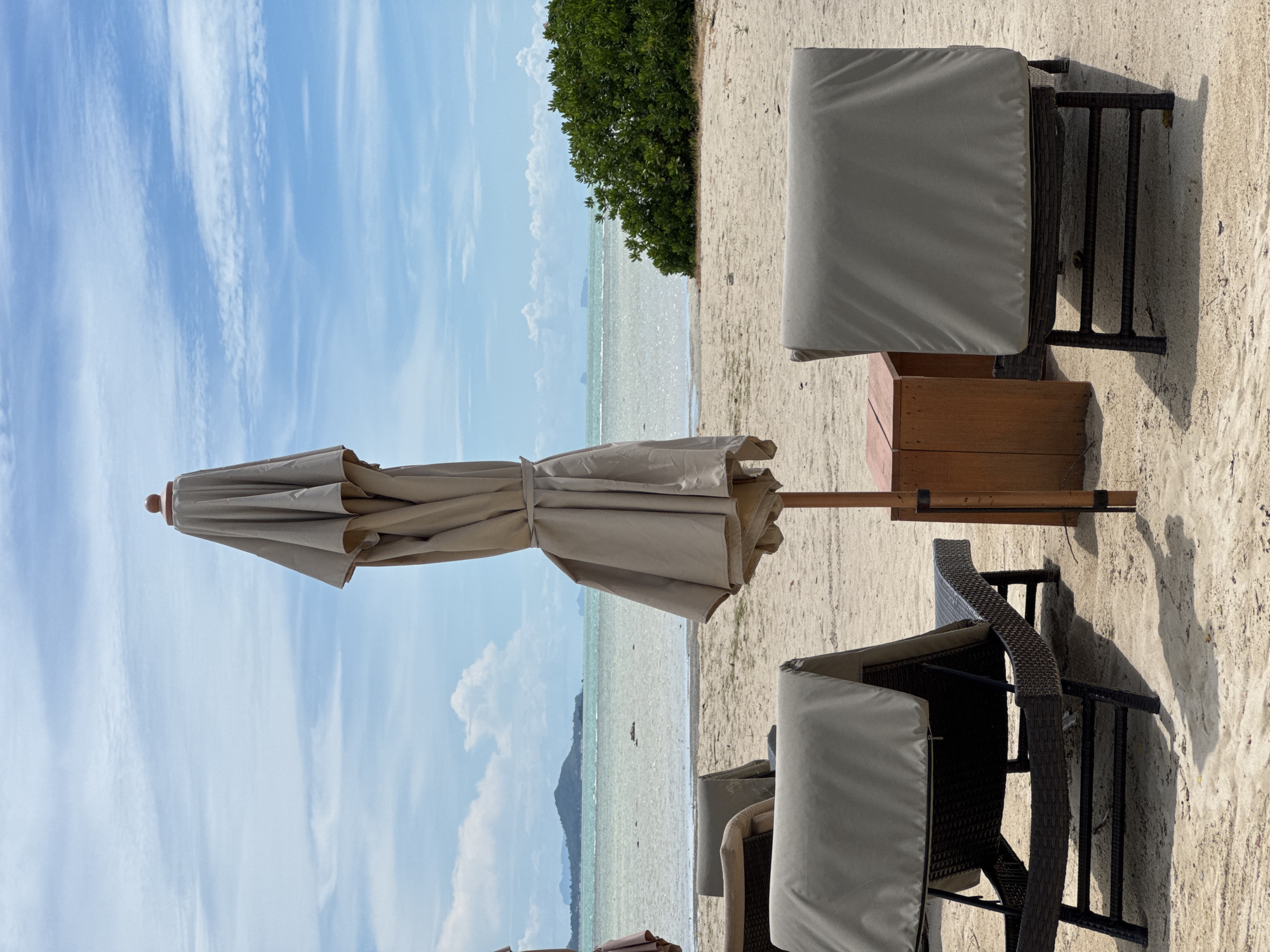 The image captures a serene beach scene in Baan Plai Laem, Thailand. The setting is a sandy beach with the tranquil ocean stretching to the horizon. The sky is a clear blue, dotted with wispy white clouds, suggesting fair weather. A lush green tree stands prominently on the sand, providing a natural contrast to the pale beach and blue sky.
In the foreground, several beach chairs are covered with light grey fabric, indicating they are not in use or are being protected. A closed beach umbrella, also in a light tan color, stands nearby, its fabric neatly tied. A wooden structure, possibly a small table or part of the umbrella stand, is visible. The sunlight casts distinct shadows on the sand, suggesting it is daytime, likely late morning or early afternoon. The overall mood is one of peaceful relaxation and quietude, typical of a tropical beach setting. There is no visible text in the image.