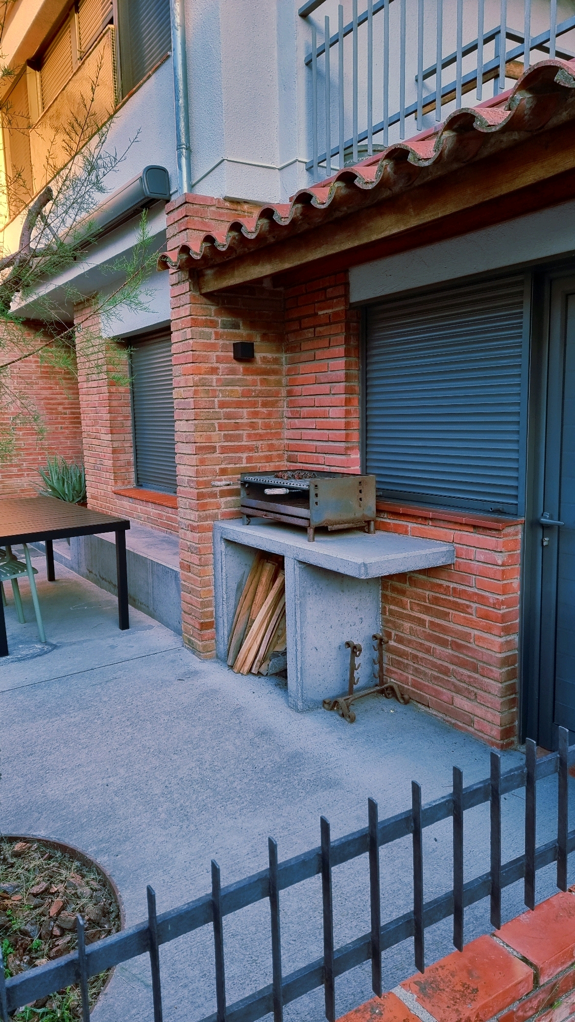 This is an outdoor scene in Sant Joan Les Fonts, Spain, featuring a brick exterior wall of a building. In the foreground, a grey concrete patio is visible, with a black metal fence running across the bottom. To the left, a dark rectangular table with a black metal frame and a dark tabletop stands on the patio, with a green chair partially visible underneath. To the right, a concrete barbecue area is built into the wall. The barbecue unit itself is a metal grill with a handle and a metal tray underneath, containing charcoal. Stacked firewood is stored within the open base of the concrete barbecue structure. The wall behind the barbecue has a grey roller shutter window. Above the barbecue and window, there is a red tiled roof overhang supported by brickwork. A tree branch extends into the frame from the left side. A metal drainpipe runs vertically down the exterior wall.