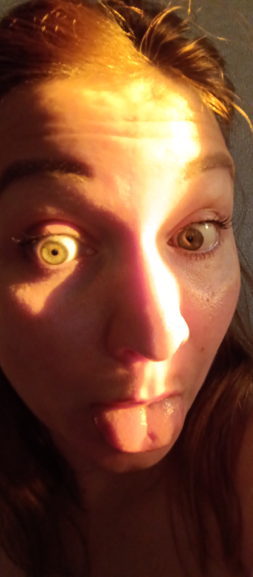 A close-up selfie shows a light-skinned woman sticking her tongue out. Her left eye is illuminated, appearing yellow, while her right eye is brown and slightly unfocused. Strong light casts shadows across her face, highlighting her nose and forehead. The location is not discernible from the image.