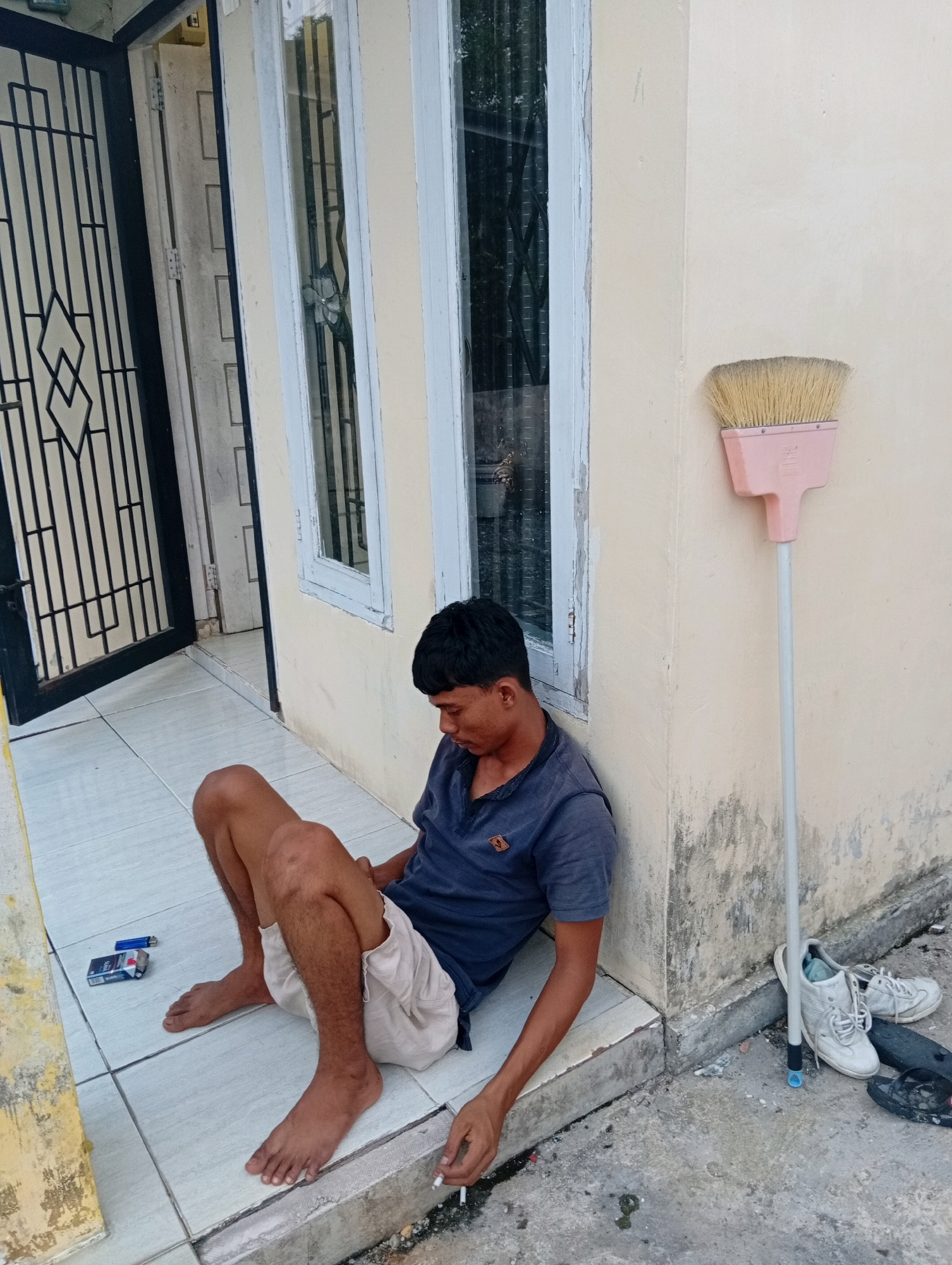 In Kualu, Indonesia, a young man sits on the tiled floor against a light-colored wall, leaning his head down. He is wearing a dark blue polo shirt and light-colored shorts. His legs are crossed, and his feet are bare. A pack of cigarettes and a lighter are placed on the floor to his left. He is holding a lit cigarette in his right hand, with the ash dangling. To his right, a broom with a pink head and white handle stands upright against the wall. A pair of white sneakers and a pair of black flip-flops are on the ground near the broom. Behind him, a black metal gate is partially visible, and a window with white trim is on the wall. The environment appears to be an outdoor entryway or porch area.
