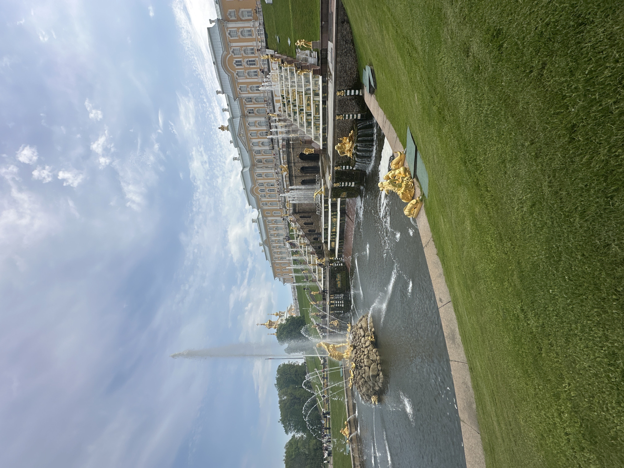 The image depicts the Grand Cascade, a prominent fountain complex in Peterhof, Russia. The scene captures the Lower Gardens and the Grand Palace in the background. The Grand Cascade consists of a series of gilded statues, cascading waterfalls, and fountains. A tall jet of water shoots upwards from the center of the main fountain. The landscape includes manicured lawns and trees. The sky is partly cloudy. No people are clearly visible in the image, though some may be obscured by the trees and distance.