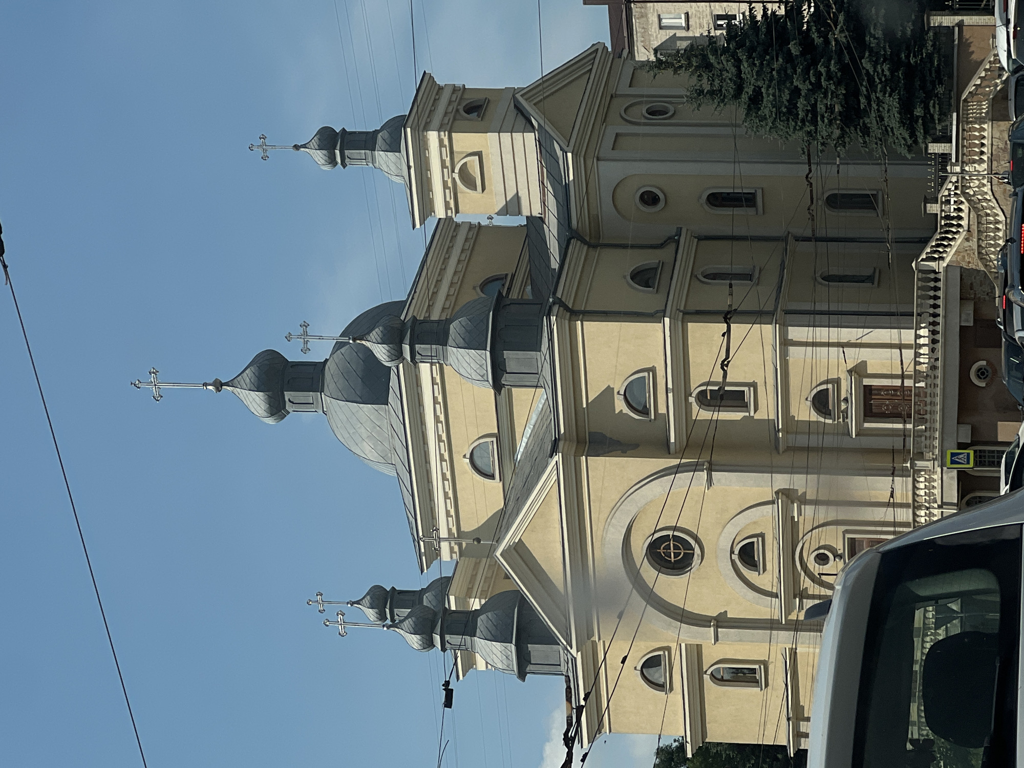 The image shows the Ternopil Theological Seminary, a religious educational institution located in Ternopil, Ukraine. The seminary is housed in a striking yellow building with a prominent dome and several smaller cupolas, each topped with a cross. The architecture features classical elements, with arched windows and decorative cornices. Electrical wires crisscross the sky in the foreground, suggesting an urban setting. A tall evergreen tree stands to the right of the building. In the lower part of the image, the roof and windshield of a silver vehicle are visible, indicating that the photo was taken from a car. The sky is clear and blue, with a few wispy clouds. The scene is a snapshot of the seminary under a bright, sunny sky.
