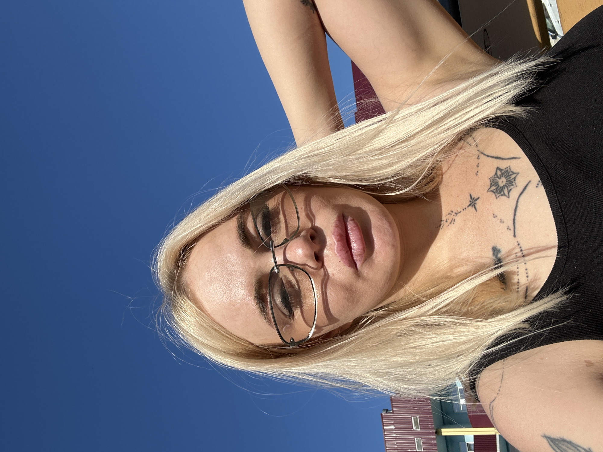 A woman with blonde hair and glasses is taking a selfie outdoors. She is wearing a black tank top and has tattoos on her chest and arm. The background shows a clear blue sky and a building with red siding. The lighting suggests it is a sunny day.