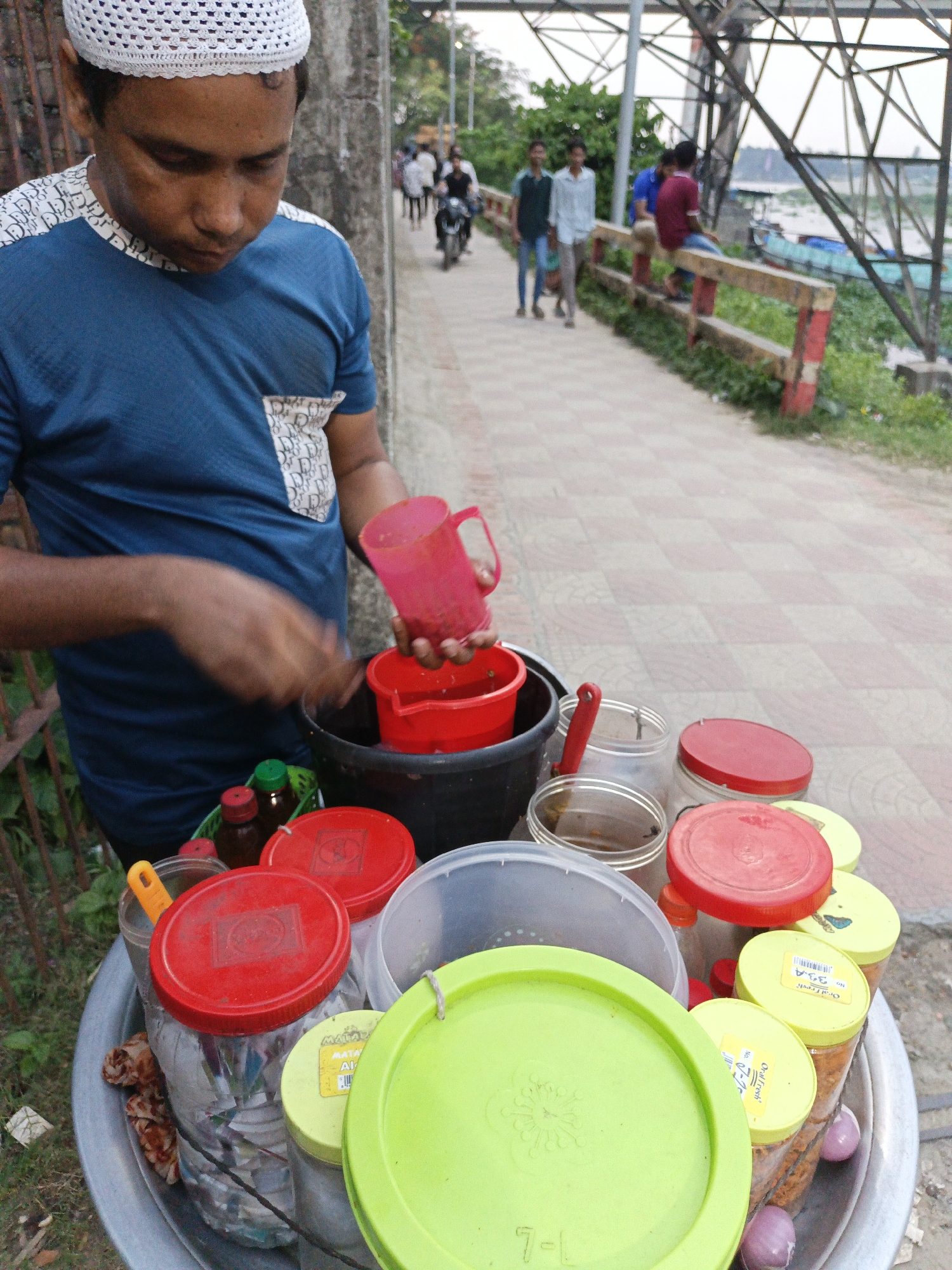 A man in a blue shirt and white crocheted cap is preparing food from a mobile cart on a paved walkway. He is pouring liquid from a pink pitcher into a red container. The cart is filled with various containers, mostly plastic, with lids in red, green, and yellow. Some containers are labeled "Oral Fresh" and "MATAR." In the background, several people are walking along the walkway, and one person is riding a motorcycle. There is also a metal structure resembling a bridge or power lines in the background. Vegetation and a river can also be seen in the distance.