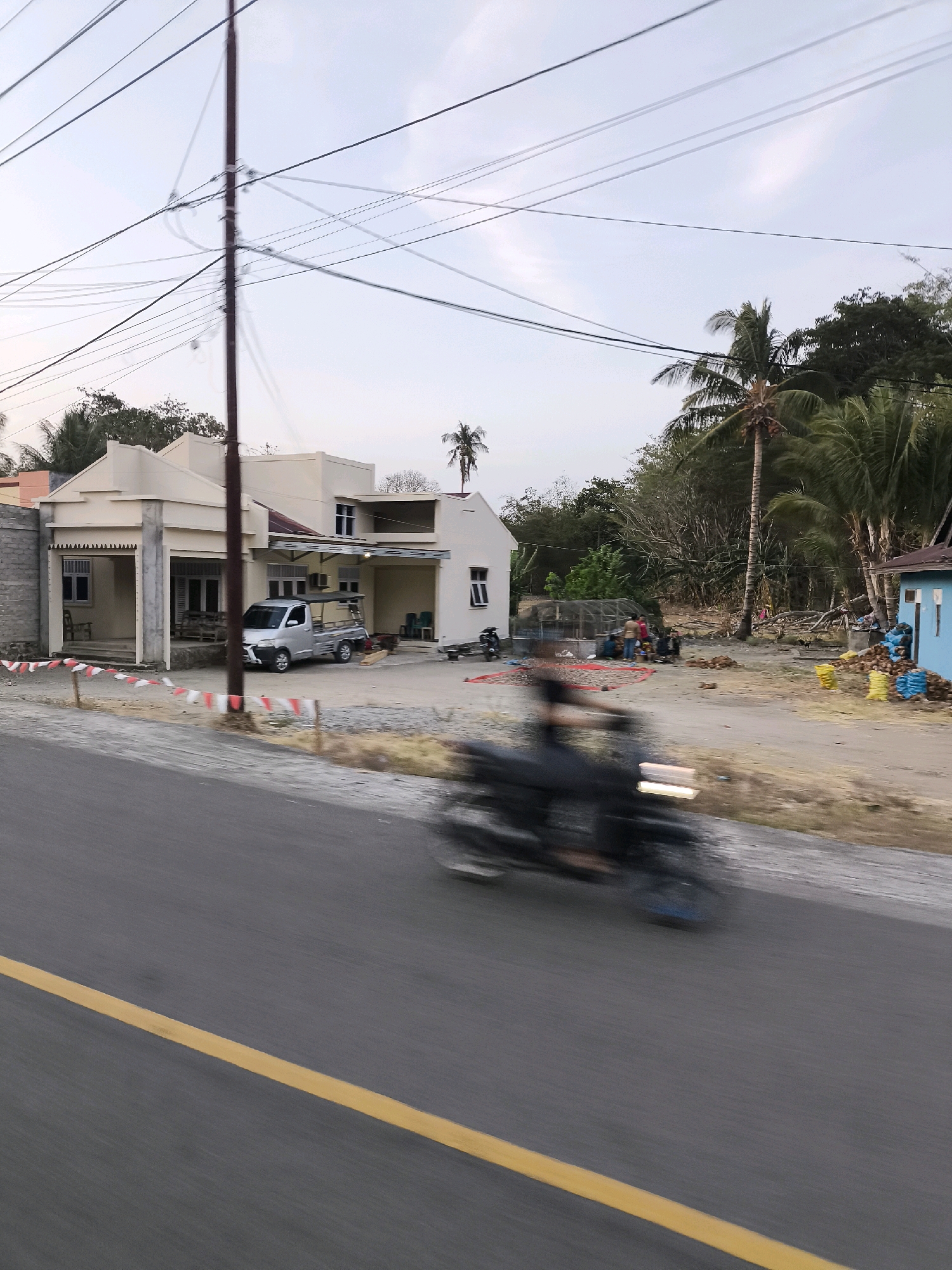 The image captures a street scene in a rural area of Indonesia. A paved road with a yellow line runs diagonally across the foreground. A motorcycle is in motion on the road, appearing blurred due to its speed. On the right side of the road, there are two light blue plastic bags and a pile of coconuts, and a light blue building. Further in the background, a cluster of buildings includes a pale yellow house with a covered porch supported by columns and a silver utility truck parked in front of it. There is also a dark-colored motorcycle parked near a white building. Several people are visible near the white building, some standing and some sitting on a red mat, with what appears to be dried produce laid out. The environment is characterized by tropical vegetation, including palm trees, and a background of dense foliage. Utility poles and overhead wires are prominent in the sky.