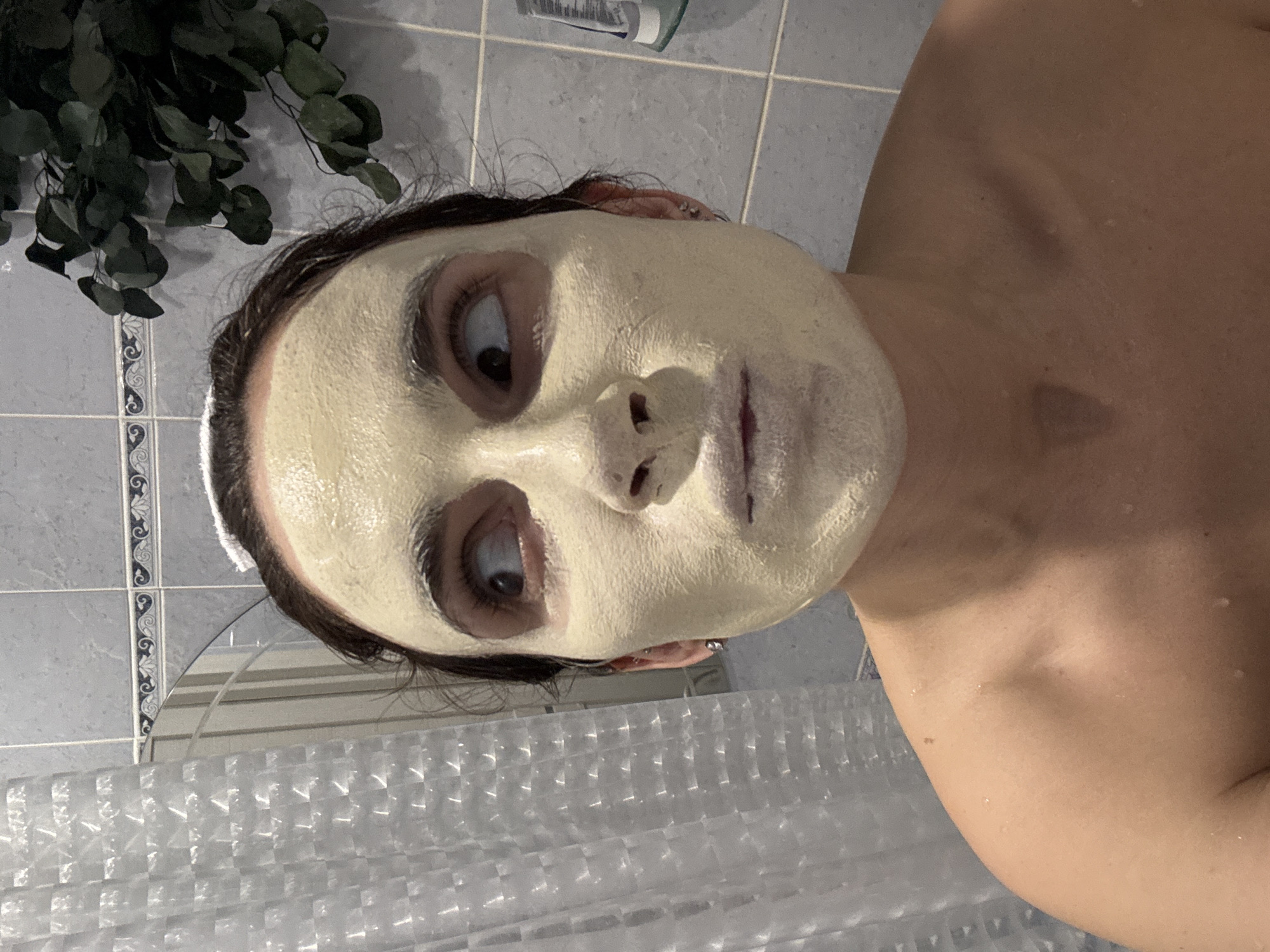 A woman with a light-yellow facial mask is in a bathroom with light-blue and white tiles. She has dark hair and a white towel headband. A plant is in the upper left corner of the frame. A shower curtain is visible in the lower part of the image. There is no evidence to suggest this image was taken in the city of Chaikovsky, Russia.
