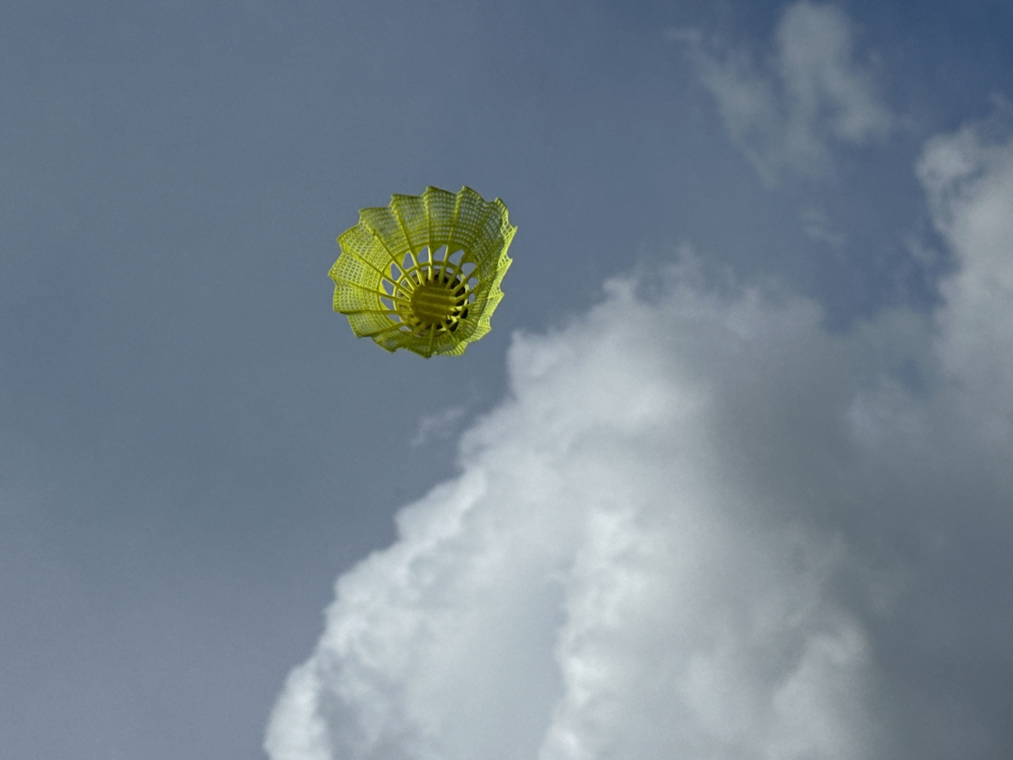 A yellow shuttlecock is suspended in the air against a backdrop of gray sky and white clouds. No people are visible in the scene. The location is not possible to determine from the image.
