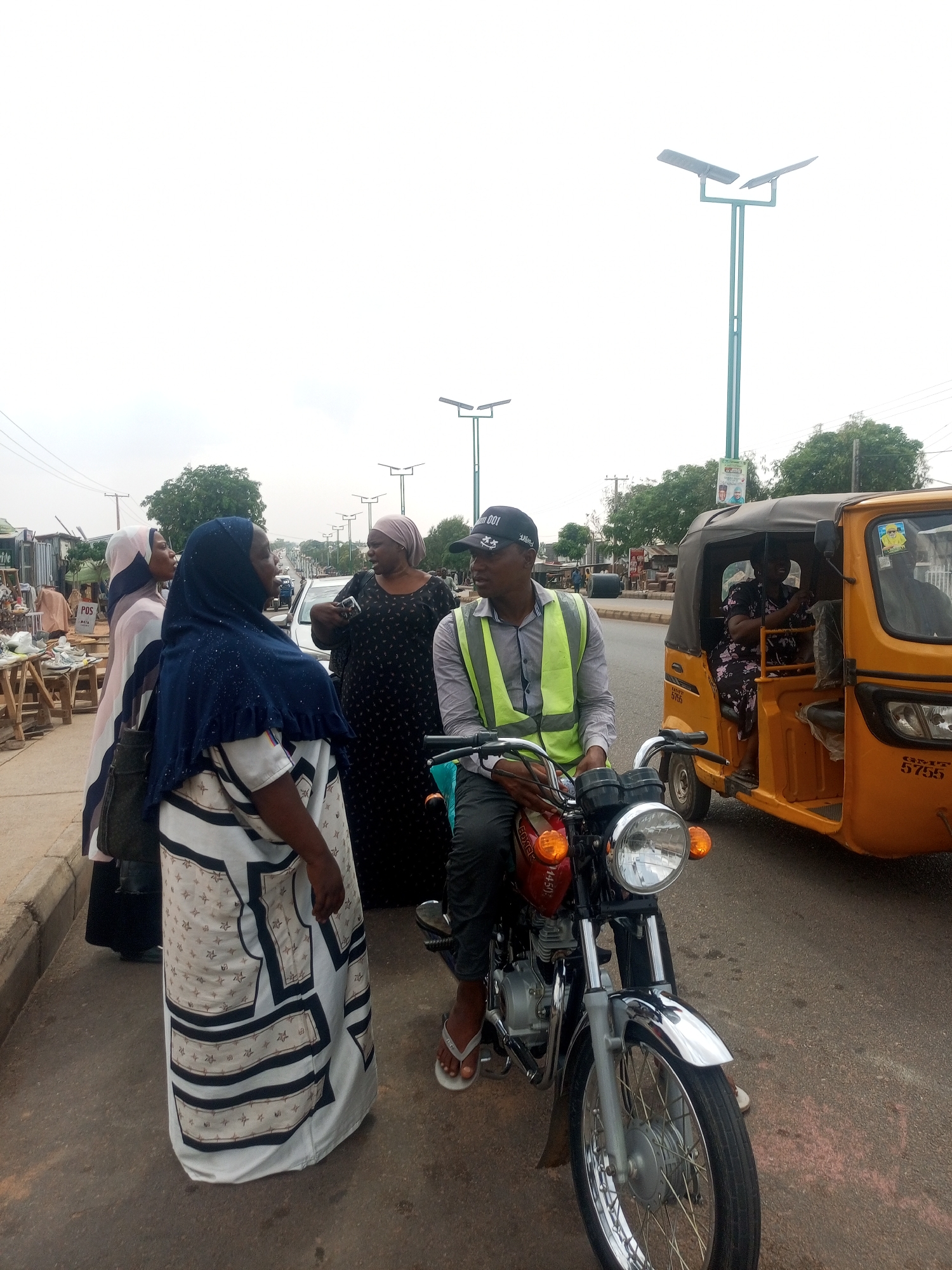 A street scene depicts a group of four women standing on a sidewalk, engaged in conversation with a man seated on a red motorcycle. The women are wearing modest clothing, with two donning headscarves or hijabs. The man on the motorcycle is wearing a neon yellow vest and a dark baseball cap. Behind them is a yellow three-wheeled vehicle, with two individuals visible inside. The street is paved and leads to a more distant part of the city with buildings and vehicles. Tall streetlights line the road. The background suggests a populated area, likely a market or business district.
