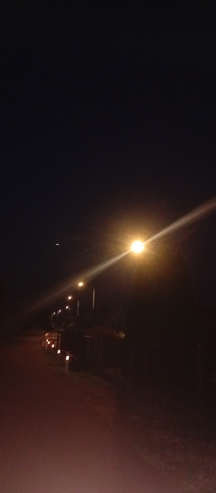 The image depicts a dark, rural road in Dolní Marklovice, Czechia, illuminated by streetlights. The perspective is from ground level, looking down a paved road bordered by low-lying lights. Several streetlights are visible along the right side of the road, casting a bright yellow glow and creating light beams that extend across the scene. The sky is a deep, dark blue, indicating nighttime. The environment appears to be sparsely populated, with no people or vehicles present. The overall atmosphere is quiet and serene, characteristic of a rural setting at night.