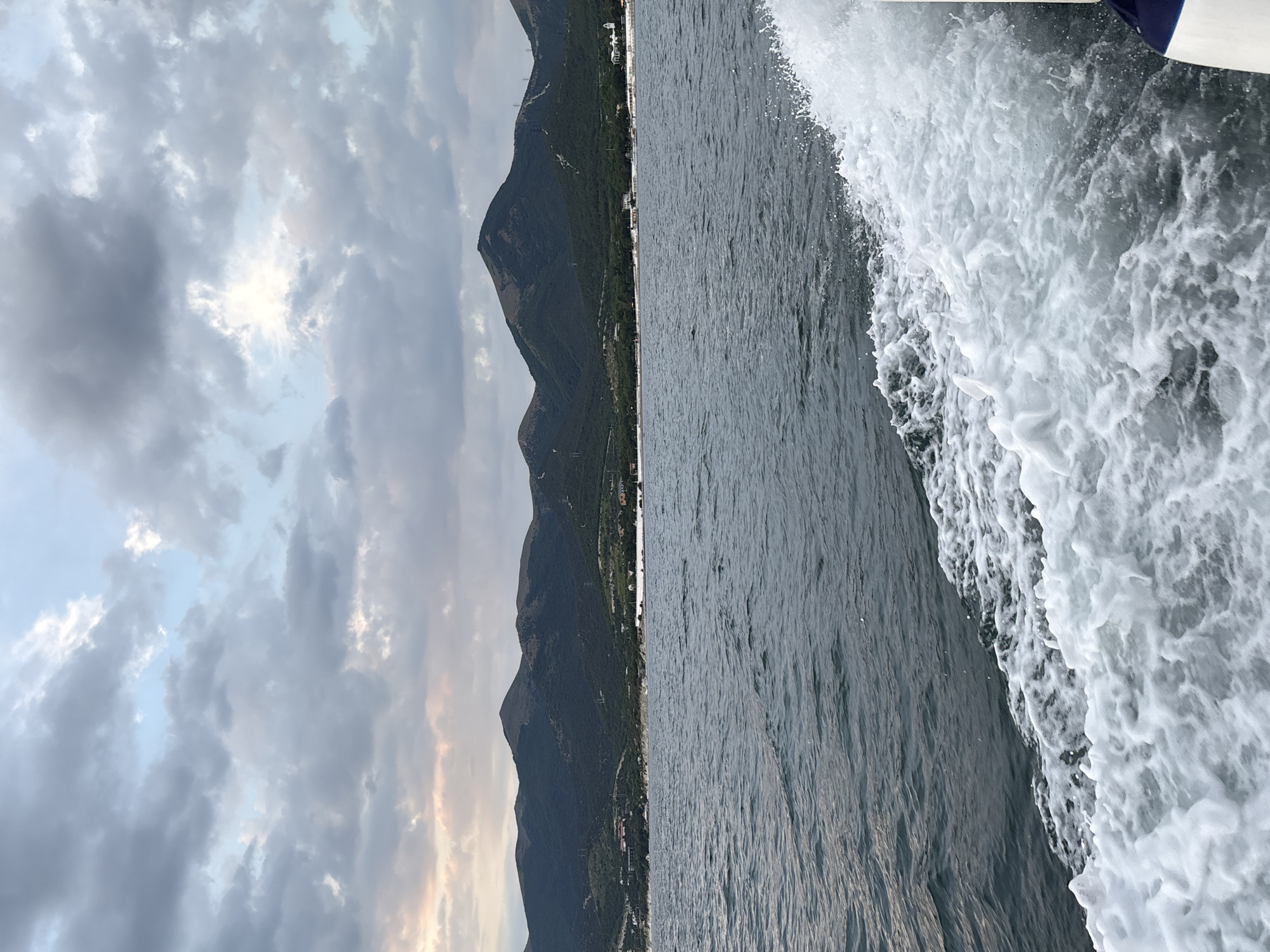 The image shows a view from a boat moving on the Black Sea, with the coastline of Kabardinka, Russia, in the background. The sea is choppy, with white foam trailing from the boat. In the distance, a range of green, tree-covered mountains forms the coastline, with a few buildings visible nestled at their base. The sky is overcast with dramatic clouds, tinged with the soft colors of either sunrise or sunset. No people or specific landmarks are clearly identifiable in this scene.