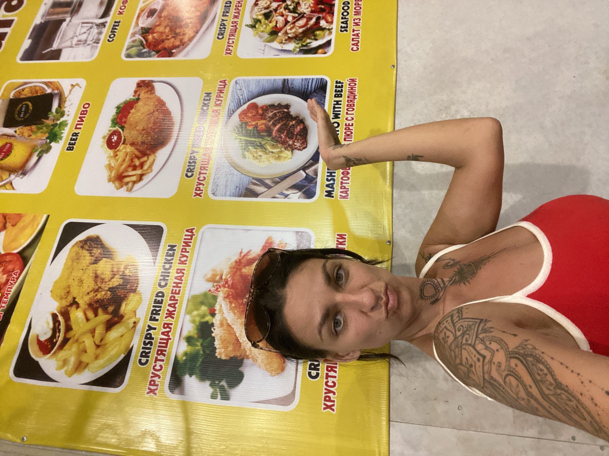 This is a selfie taken outdoors, likely in Phan Thiết, Vietnam, as indicated by the presence of Vietnamese text on the menu. A woman with dark hair and sunglasses pushed up on her head is lying down, looking at the camera with a neutral expression. She is wearing a red top with white trim. She has extensive black tattoos on her arms and neck.
She is lying next to a large, yellow banner that appears to be a menu for a food establishment. The menu features pictures of various dishes, including crispy fried chicken, beef with mashed potatoes, seafood salad, and beer. Some of the text on the menu is in English, and some is in Russian, and it appears to have some Vietnamese text as well. The banner is positioned horizontally in the frame, with the woman lying beside it. The background is a light-colored, textured surface that resembles concrete or stone. The lighting suggests it is daytime. The overall scene suggests a casual, possibly tourist-oriented setting.