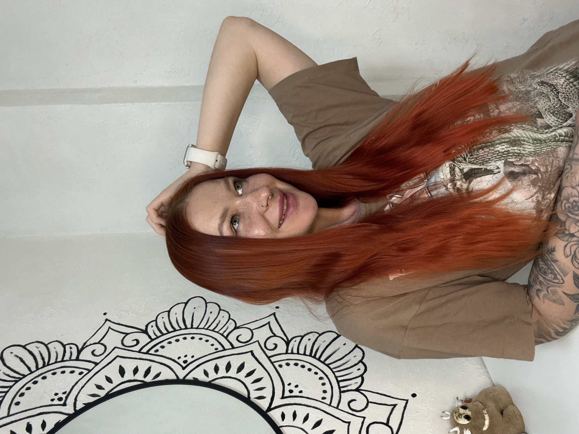 A fair-skinned woman with long, red hair poses in front of a white wall decorated with a black mandala design. The woman wears a tan-colored t-shirt with a faded design on the front and a white smartwatch on her left wrist. Tattoos are visible on her left arm. Her left hand rests on her head, and she is smiling with a nose piercing visible. A stuffed sloth toy lies on the floor next to the wall. The location is likely in a private residence, but the exact city of Rostov-on-Don, Russia, cannot be confirmed from the image alone.