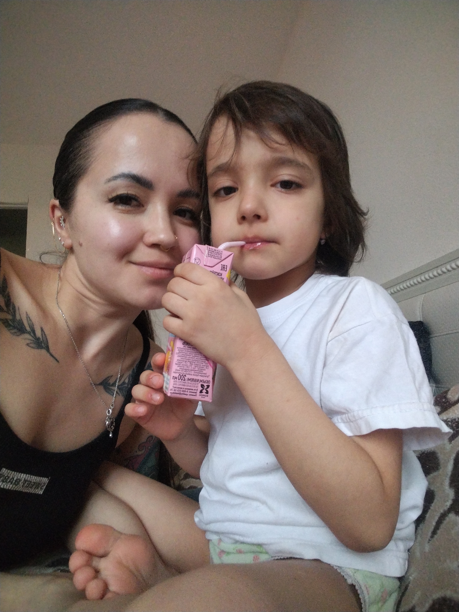 A medium shot depicts a woman and a child, likely in an indoor setting. The woman has dark hair pulled back, multiple piercings in her left ear, a nose ring, tattoos on her chest and shoulder, and a silver necklace. She is wearing a black tank top. The child, positioned to the right of the woman, has medium-length brown hair and wears a white t-shirt and a diaper. The child is holding a pink juice box with a straw in their mouth. A portion of a bed headboard is visible in the background. There is no evidence of Noviy, Russia in this image.