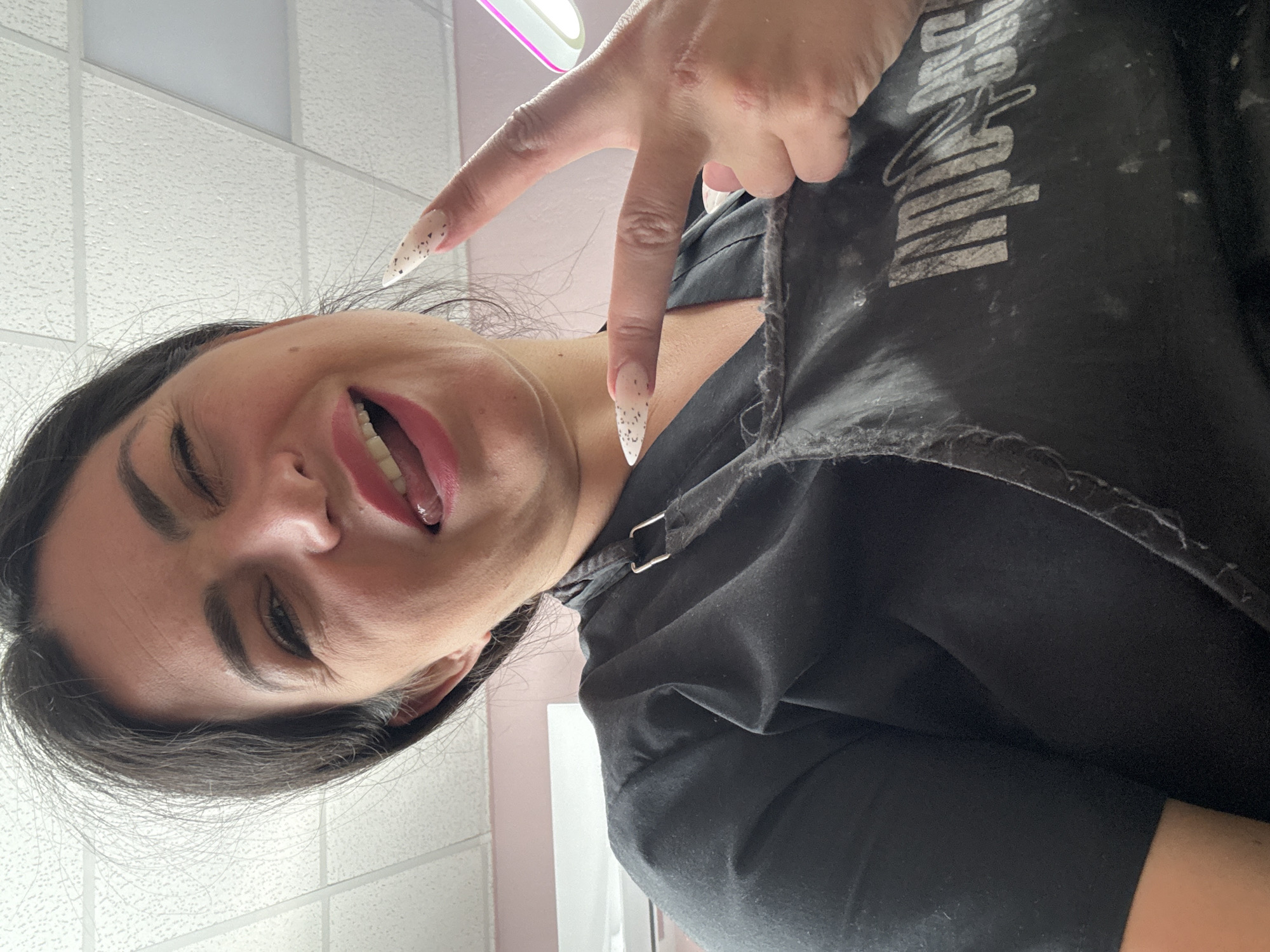 The image captures a playful moment featuring a woman posing indoors. Her face, positioned diagonally, shows her winking with her right eye and her mouth open, revealing her tongue. She has dark hair, prominent eyebrows, and wears pink lipstick. Her right hand is raised, forming a peace sign with her index and middle fingers. Her nails are long, pointed, and decorated with a light base and dark speckles.
She is wearing a black garment, likely an apron or work smock, with a visible strap and buckle near her shoulder. Distressed white text, partially reading "ROCK AND ROLL," is printed on the black fabric of the garment. The setting appears to be an indoor room, indicated by a white textured ceiling with rectangular panels in the background, a light-colored wall, and a partially visible light fixture emitting a pinkish glow. The woman's expression suggests a cheerful and confident mood.
