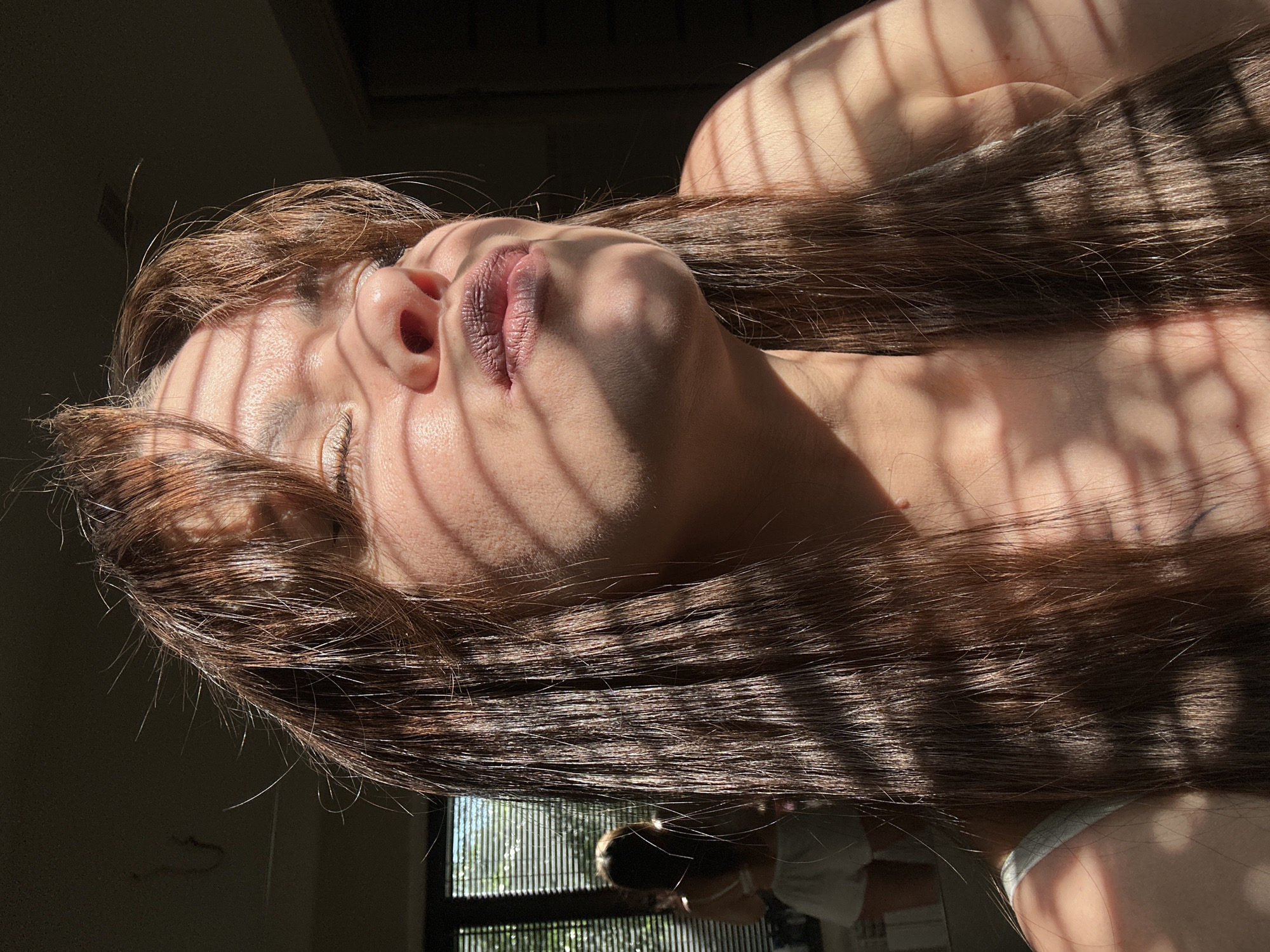 The image depicts a close-up of a young woman's face, bathed in dappled sunlight filtered through blinds. She has long, dark brown hair that falls around her shoulders and face. Her eyes are closed, and her lips are pursed in a kiss. The lighting creates a striking contrast between light and shadow across her features, highlighting her skin and hair. In the background, a reflection in a mirror shows another person, also with long hair, wearing a white top and shorts. The setting appears to be indoors, with a window and blinds visible, suggesting a residential environment. The image does not provide any specific information that would allow for localization to Chaikovsky, Russia.