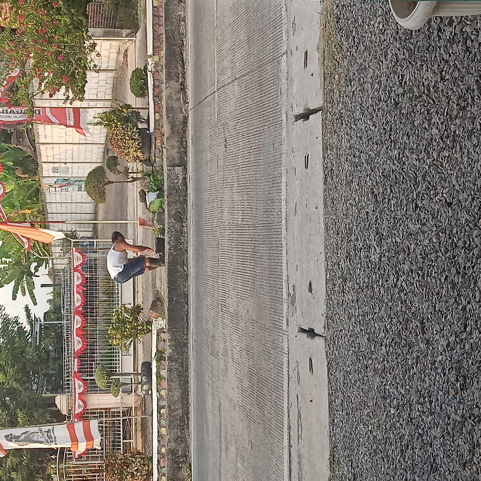 In Plaosan, Indonesia, a young man wearing a white tank top and blue shorts, with dark boots, is bent over, possibly cleaning or tending to something on the edge of a paved walkway. He is positioned near a black bucket. The background features a white brick wall, a metal fence decorated with red and white pennant banners, and flags. Lush green trees and banana plants are visible, along with decorative potted plants. The scene is set alongside a paved road with a concrete divider. The foreground consists of a gravel-covered area and a concrete drainage channel.