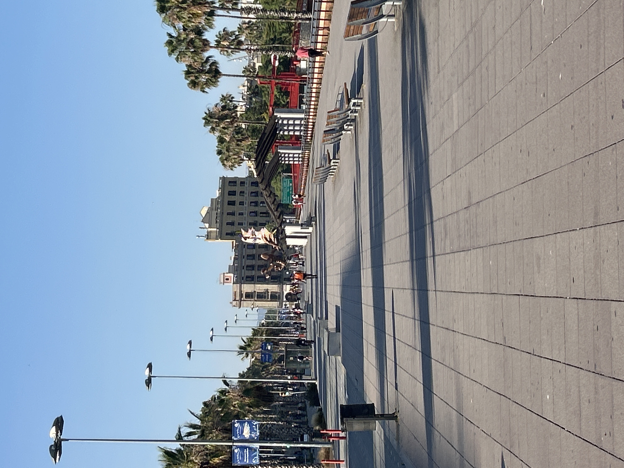 This media file depicts a sunny day in Barcelona, Spain, along a wide, paved promenade lined with palm trees. The promenade is populated by people walking and sitting on benches. The scene appears to be a lively public space, possibly a boardwalk or park, with several modern buildings visible in the background.
The primary subjects include numerous palm trees, a paved walkway with distinct horizontal lines, and several benches positioned along the promenade. People of various ages are present, some strolling casually, others seated. The architectural style of the buildings suggests a European urban setting, with a mix of contemporary and older structures. Streetlights with a distinctive design are visible, indicating a public area designed for pedestrian use.
The activity taking place is predominantly leisure and recreation. People are enjoying the outdoors, walking, and relaxing. The presence of benches suggests a place for rest and observation. The clear blue sky and bright sunlight indicate pleasant weather conditions, likely mid-morning or afternoon given the length and angle of the shadows cast by the palm trees and benches.
Notable details include the repeating pattern of the paving stones and the linear arrangement of the benches, which creates a sense of order and depth. The palm trees add a tropical or Mediterranean feel to the urban environment. There are some blue signs with white text visible, but the text is illegible in the provided image. The overall mood is relaxed and serene, characteristic of a pleasant day in a popular public space.
