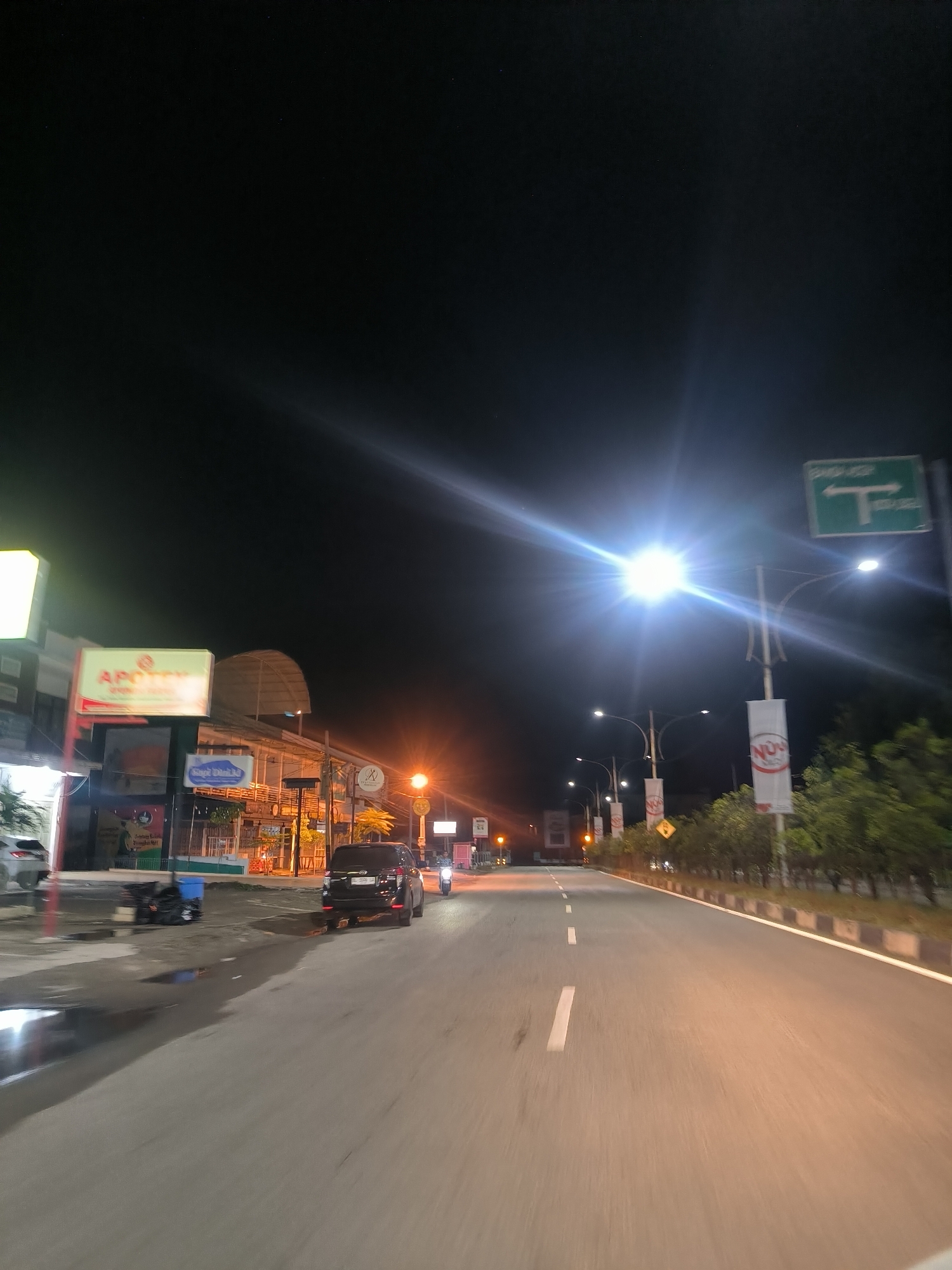 The image shows a nighttime street scene in the city of Sigli, Indonesia, likely a commercial area. A paved road with white lane markings occupies the foreground, with a slight sheen of water on the surface. Several vehicles are present: a dark SUV is parked or stopped on the right side of the road, and a motorcycle is seen in the distance to the right. On the left side of the road, buildings with illuminated signs are visible. One sign reads "APOTEK" (pharmacy) in red and white. A street light with a bright lens flare illuminates the scene from above and to the right. Several other streetlights and decorative banners are also visible. Trees and shrubbery line the right side of the road. A green traffic sign with a white "T" symbol indicating a junction is mounted on a pole. The overall atmosphere is one of a moderately busy urban street at night.