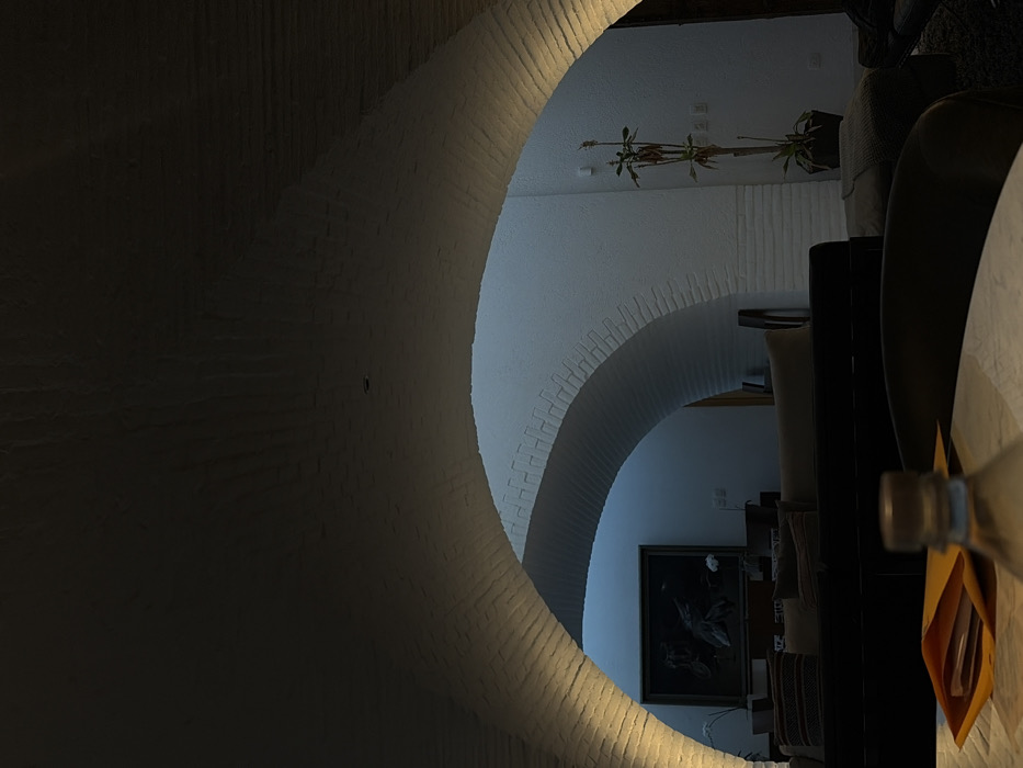 The image showcases a layered perspective through a series of archways. The arches are made of white bricks and progressively lead into the interior space. The background reveals a glimpse of a framed painting and a plant suspended from the ceiling. A partially visible table and chairs occupy the foreground, with a bottle and envelope positioned at the edge. The light source appears to originate from the archways, casting shadows and highlights on the brickwork.
