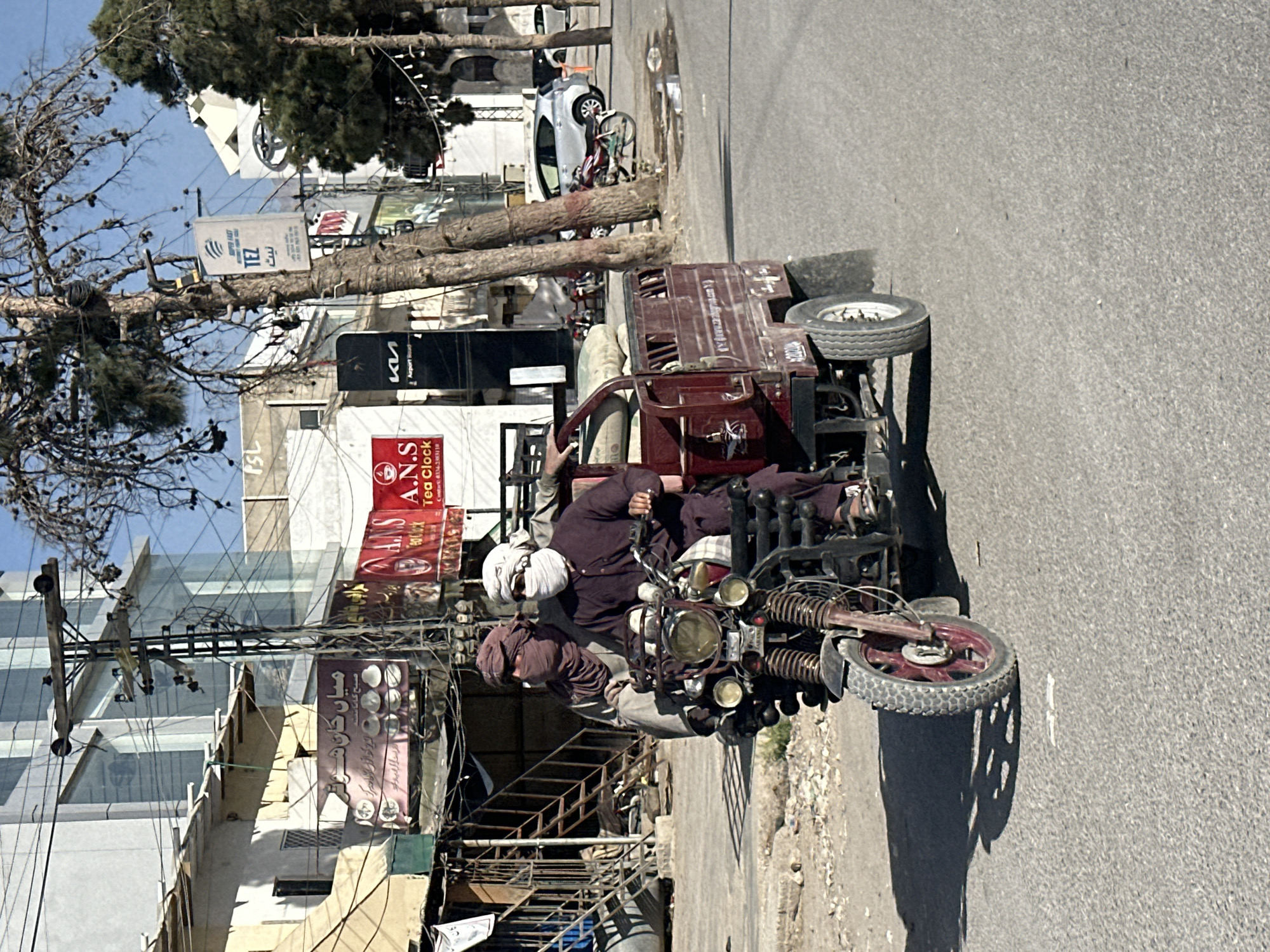 An outdoor, rotated image depicts a street scene with two men on a motorized three-wheeler, commonly known as an auto-rickshaw or rickshaw, on a sunny day. The vehicle, a maroon color with a cargo bed at the back, is in motion on an asphalt road. One man, wearing a white turban and dark clothing, is driving, holding the handlebars. Another man, wearing a brown head covering, is seated as a passenger behind him.
In the background, there are multi-story buildings and numerous power lines strung between poles. Large trees with green foliage are visible, casting shadows on the ground. A white car and a bicycle are partially visible further down the street. The sky is clear blue.
Several signs are visible on the buildings, primarily featuring text in Urdu/Arabic script and some English. Visible text includes "ميان كان هوت" (Mian Kan Hot), "صبح گالانت" (Subah Gallant), "PSL", "SUPER FAST", "TEZ", "A.N.S Tea Clock" with a contact number, "ки Airport Road" (likely Kia on Airport Road), and "نيت" (Net). Another sign shows "1900" and "الحكيم" (Al-Hakim). The strong sunlight indicates it is daytime. The specific location is not identified, but the script and vehicle type suggest a South Asian or Middle Eastern setting.