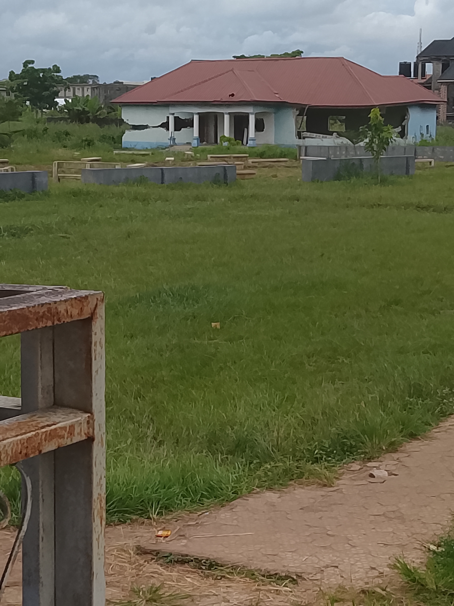 The image shows an exterior scene in Ihiagwa, Nigeria, under an overcast sky, suggesting it might be daytime with cloudy weather. The focal point is a partially constructed or dilapidated building with a red roof and light blue walls. The building appears to have suffered damage, with large sections of the walls missing, revealing the interior structure. Several white columns are visible along the front of the building. In front of the house, there are concrete boundary walls and some wooden planks arranged in the grass, possibly indicating ongoing construction or foundation work. The foreground features a grassy field and a section of a paved path. On the left side of the image, a rusty metal structure, possibly part of a gate or fence, is visible. In the background, other buildings and lush green trees can be seen, indicating a semi-urban or developing area. There are no people visible in the image, and the scene appears quiet and deserted, suggesting no activity is currently taking place. A small yellow and red object, possibly a wrapper, is visible on the paved path.