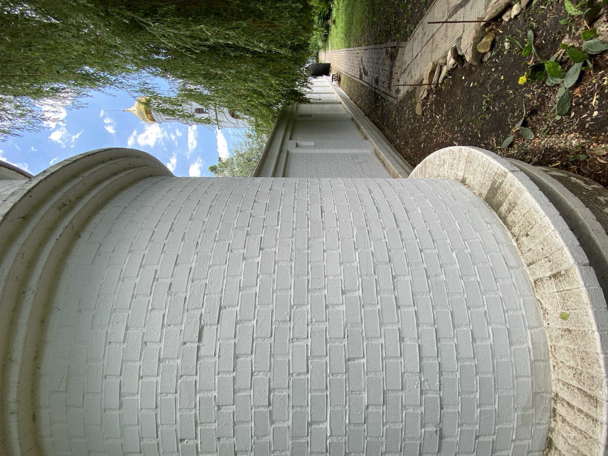 The image depicts a low-angle, sideways view of a white brick wall curving into the foreground, with a pathway receding into the distance on the right. Overhanging green foliage from a tree creates a partial canopy in the upper left portion of the frame. In the background, a white church tower with a golden dome and a spire is visible against a blue sky dotted with white clouds.
The pathway, made of light-colored rectangular stones, is bordered by dark soil and scattered rocks, with some patches of dry grass and low-lying green plants, including a prickly pear cactus with a small yellow flower. The white brick wall appears to be part of a building or structure with rounded architectural elements, showing a repeating pattern of bricks. The overall scene suggests an outdoor setting, possibly a courtyard or garden area adjacent to a religious building.
The lighting indicates it is daytime, with sunlight casting shadows and highlighting textures. The weather appears fair with a clear blue sky. No people are visible in the frame. The visual elements suggest a peaceful and serene atmosphere, typical of a quiet religious or historical site. The location context provided is Plastunovskaya, Russia.