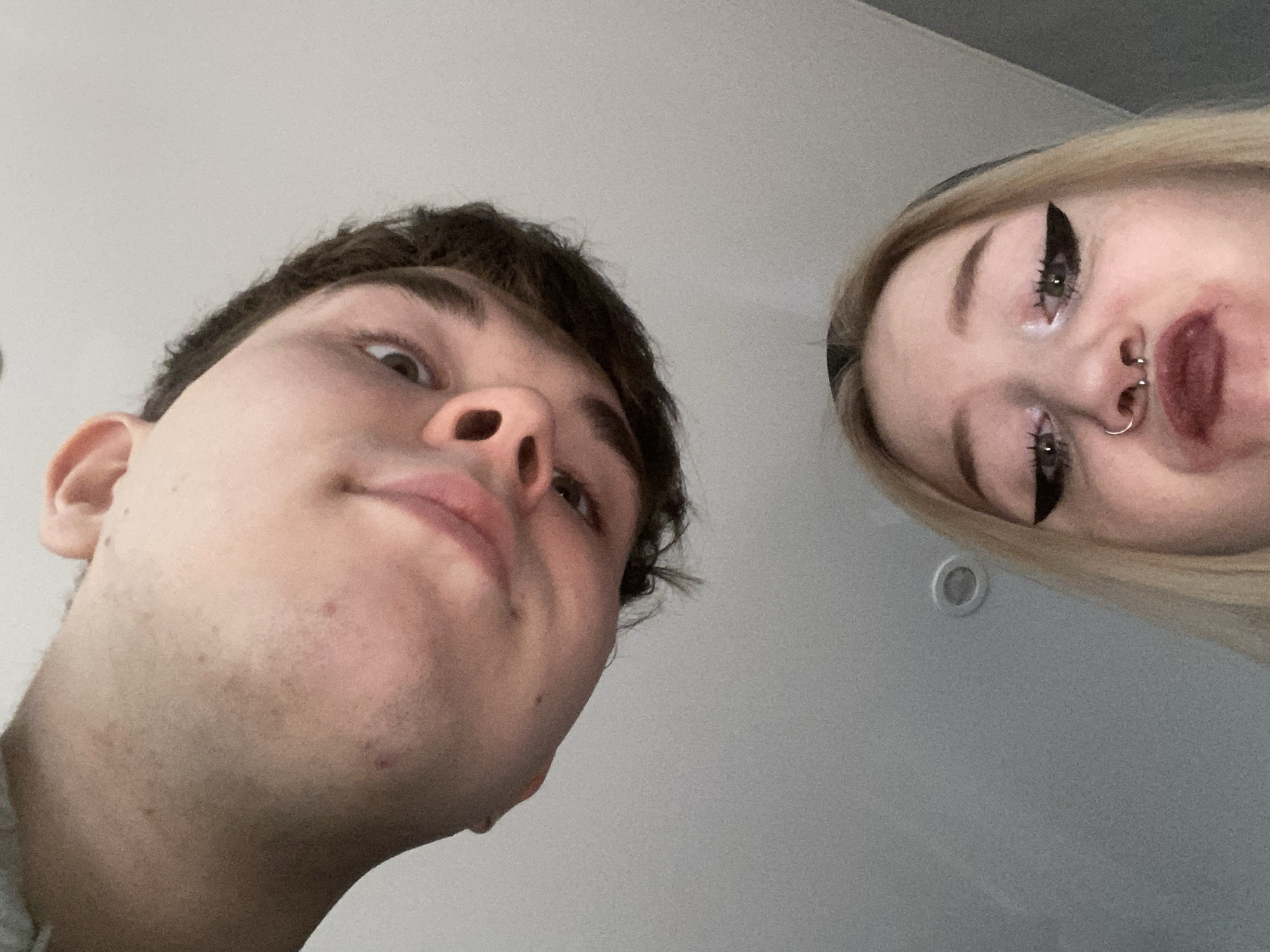A low-angle, close-up photograph depicts two individuals: a young man on the left and a young woman on the right. The man has dark hair and light skin. The woman has light blonde hair, heavy black eyeliner, and a septum piercing. The background is a light grey or white color with a small round object visible near the top center. The lighting is even and diffused. The angle suggests the picture was taken from below the subjects. There is no information in the image to suggest this photograph was taken in Izhevsk, Russia.