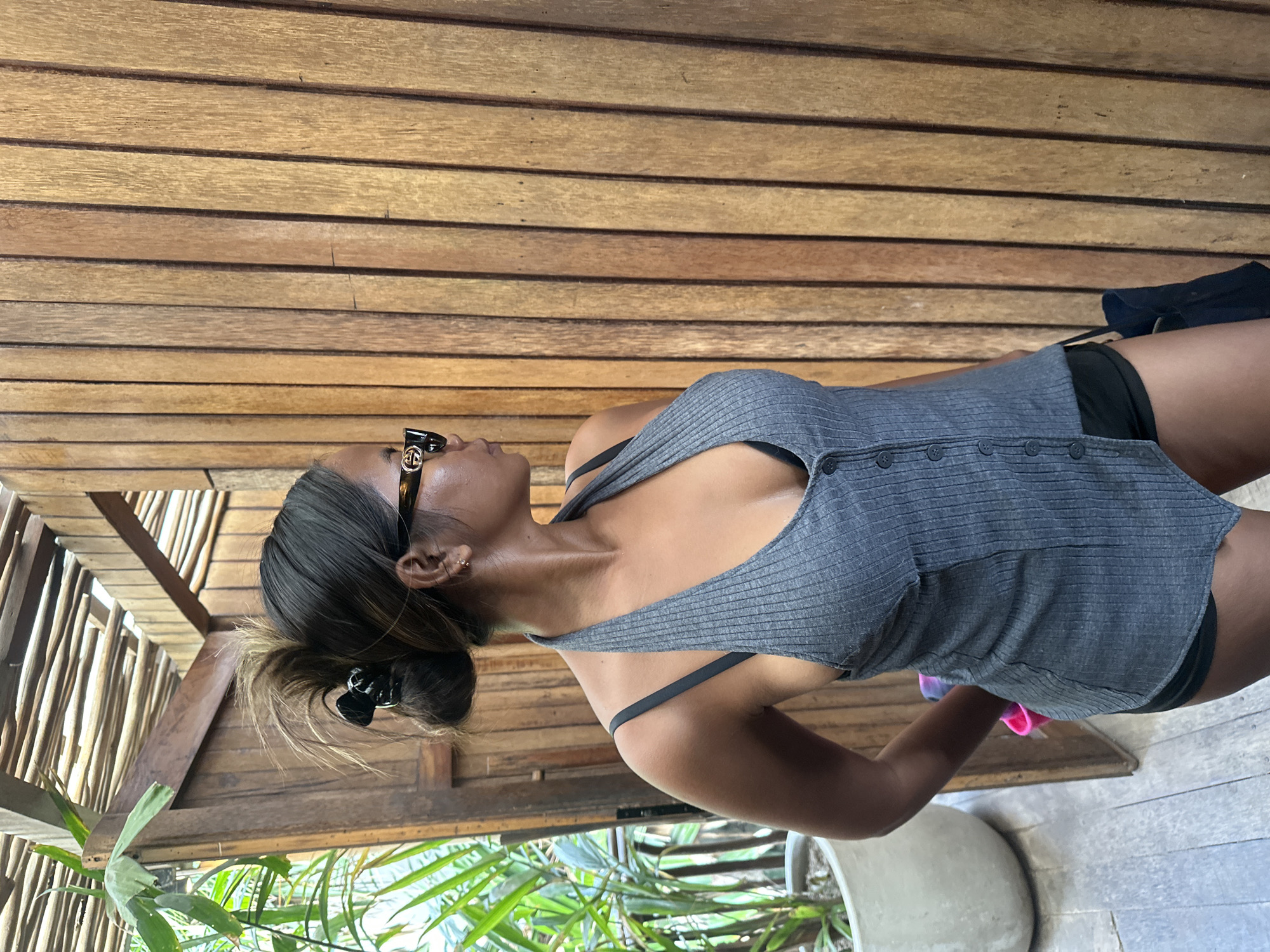 A woman stands facing right, looking off-camera, in what appears to be an outdoor or semi-outdoor area. She has her hair pulled up in a bun and is wearing sunglasses, a gray halter top, and dark shorts. Her left arm is bent, with her hand near her hip. Behind her is a wall made of vertical wooden planks, and to her left, there is a wooden doorway and lush green foliage in a large planter. The overall setting suggests a tropical or resort-like environment, possibly a bathroom or changing area, consistent with locations in North Kuta, Indonesia.