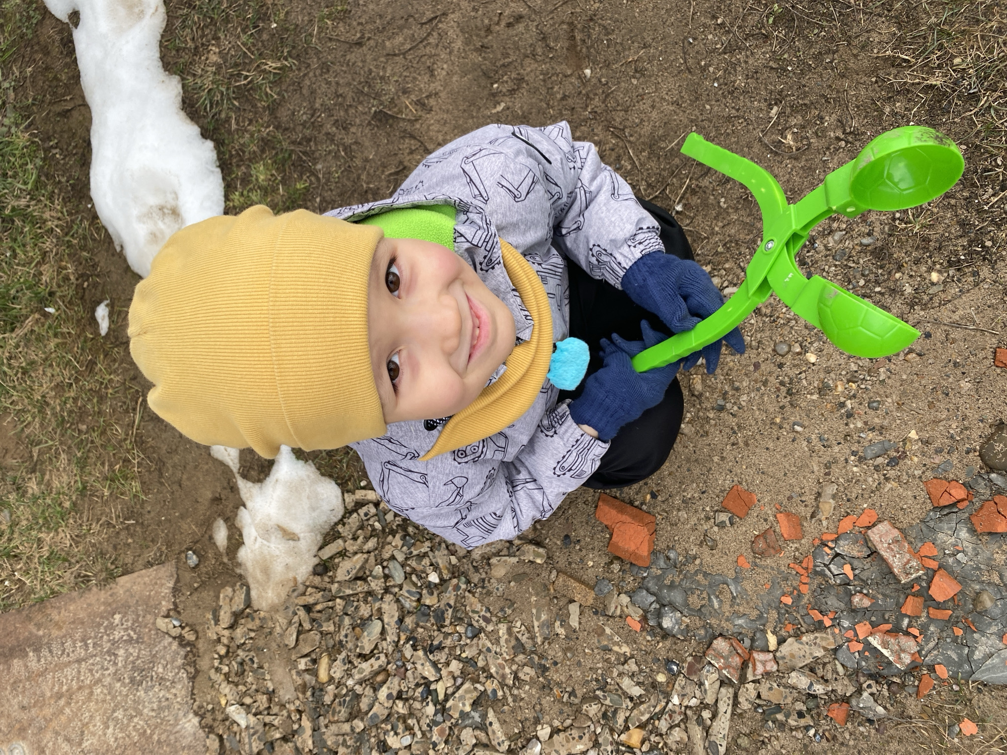 A young child wearing a yellow hat, a patterned gray jacket with a green lining, a yellow scarf, and blue gloves is crouching outdoors, holding a green plastic snow or sand tool. The child is smiling and looking up at the camera. The ground consists of dirt, gravel, and pieces of broken red brick. There is a patch of melting snow visible in the upper left corner of the frame. The photograph was taken in Chaikovsky, Russia.