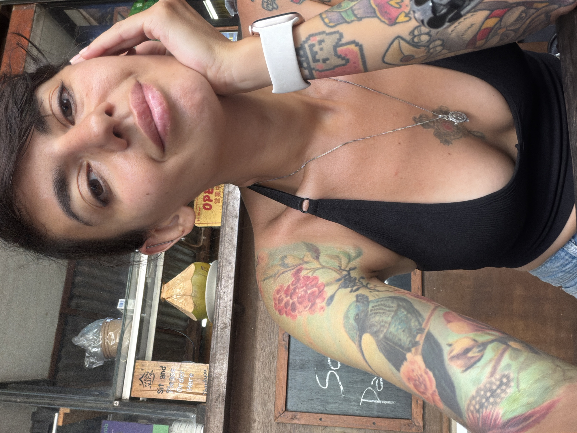 In Kuta Selatan, Indonesia, a woman with short dark hair and a tattoo on her left arm is pictured from the chest up. She is wearing a black bikini top and a silver necklace with a pendant. Her right arm is partially visible, with tattoos and a white smartwatch. In the background, there is a chalkboard with writing and a half-cut coconut. The scene appears to be outdoors, possibly at a cafe or market.