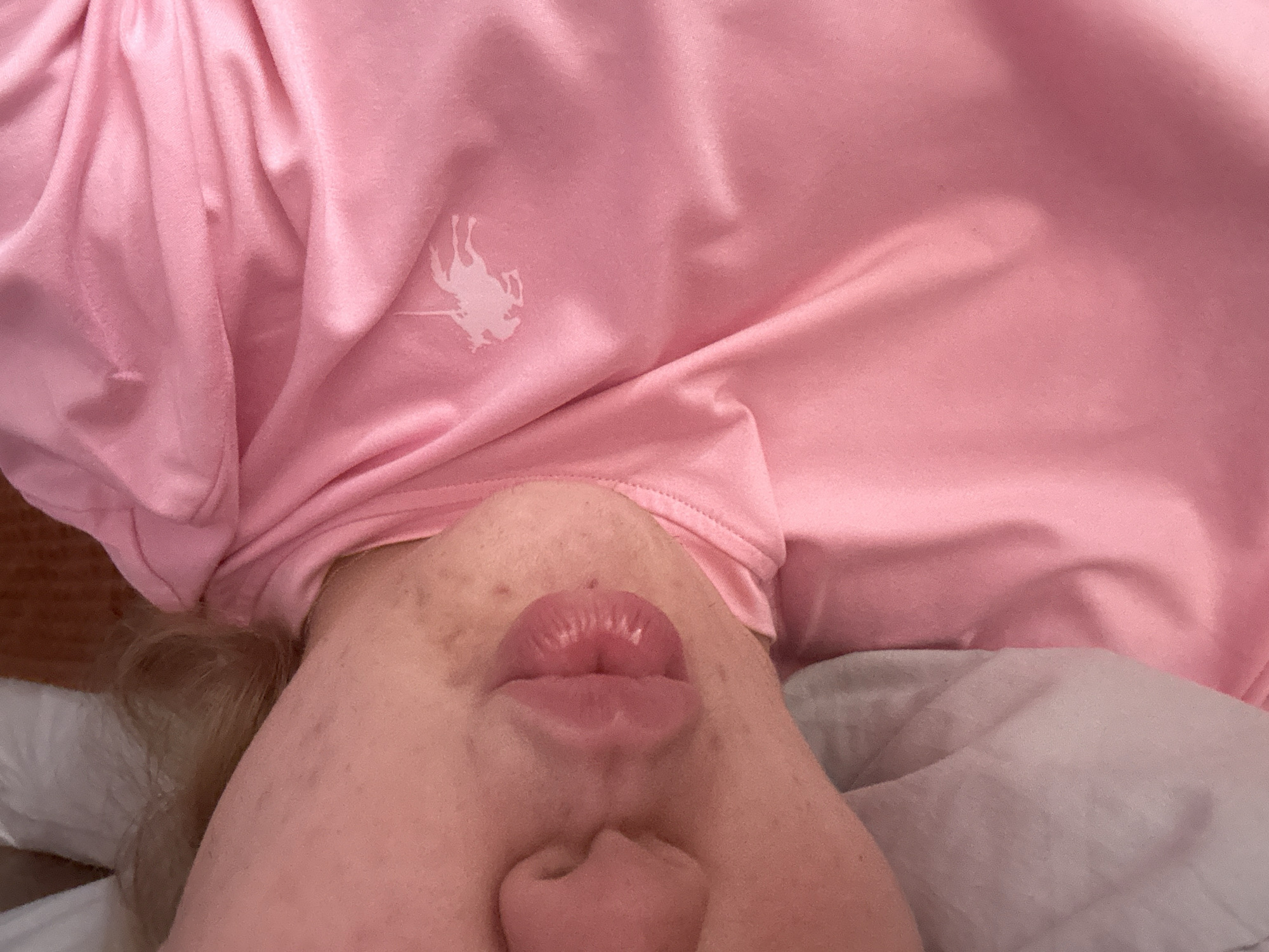 The media file is a close-up, upside-down photograph of a person's face and torso, taken from a low angle. The subject is wearing a pink, long-sleeved garment with a light-colored embroidered logo of a horse and rider on the upper chest area. The fabric of the garment is wrinkled, suggesting it might be soft or loosely fitting. The person's face is turned towards the camera, with their lips pursed as if blowing a kiss. Their skin appears fair with some freckles and blemishes. Their hair, which is light-colored and appears to be blonde, is visible near the neck area. The background consists of a light-colored surface, likely a bed or pillow, with some darker fabric visible to the left. The lighting is soft, suggesting an indoor setting, possibly during the day. The overall mood is playful and intimate. There is no visible text in the image other than the embroidered logo. The location context is Plovdiv, Bulgaria.