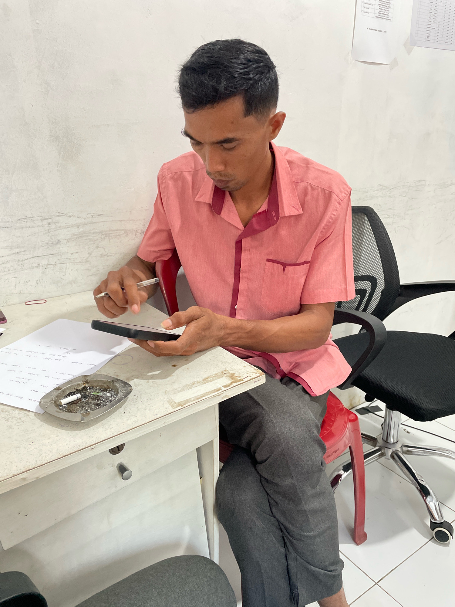 A man wearing a pink collared shirt and gray pants is sitting at a white desk in an indoor setting. He is holding a black smartphone in his left hand and a pen in his right hand, appearing to be writing on a piece of paper on the desk. To his left is a metal ashtray containing cigarette butts and ash. The desk also has a red rubber band and a portion of a document. Behind the man, a black office chair with a mesh back is visible, with part of a red plastic stool underneath. The wall behind the desk is white and textured. The scene is likely an office or a study in Kagungan Ratu, Indonesia.